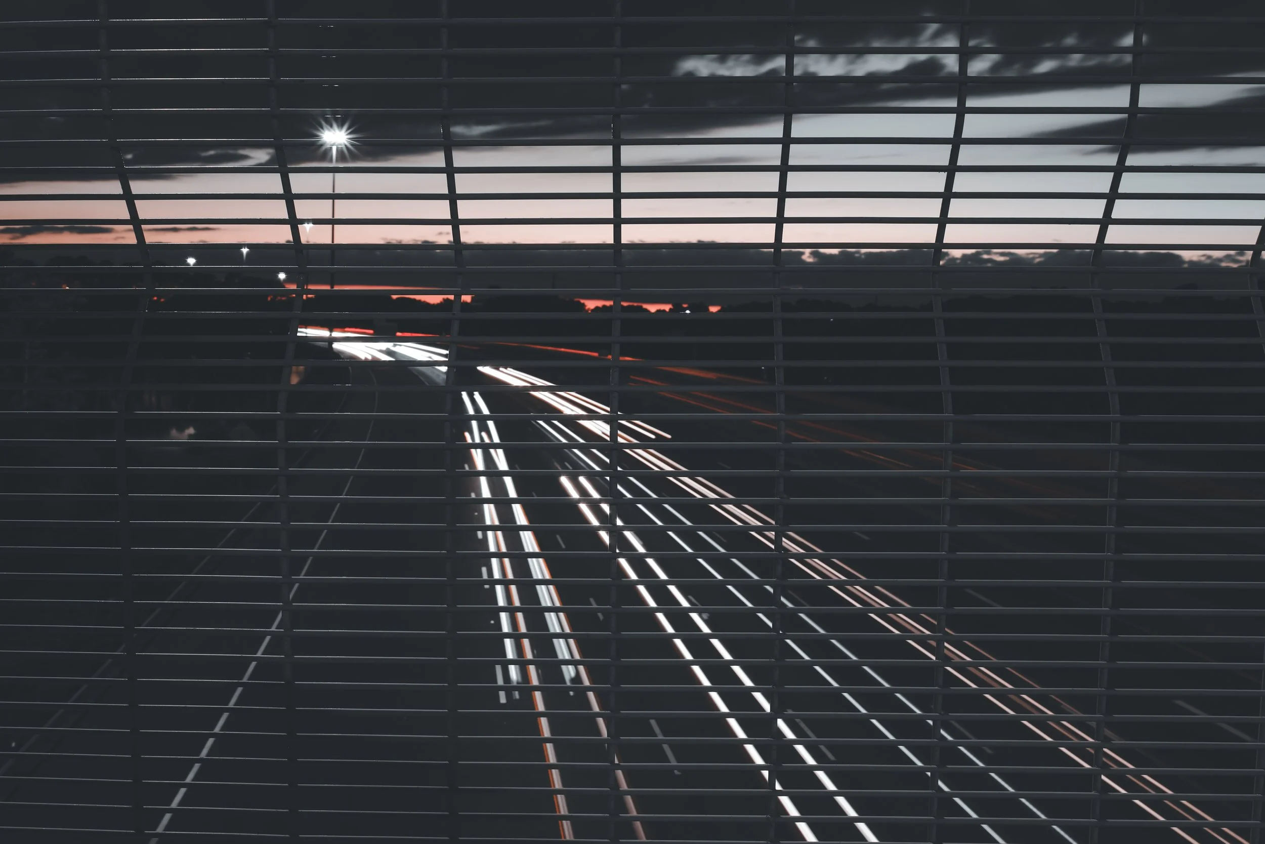 Photo of a highway at sunset viewed through horizontal metal bars, with light trails from moving vehicles.