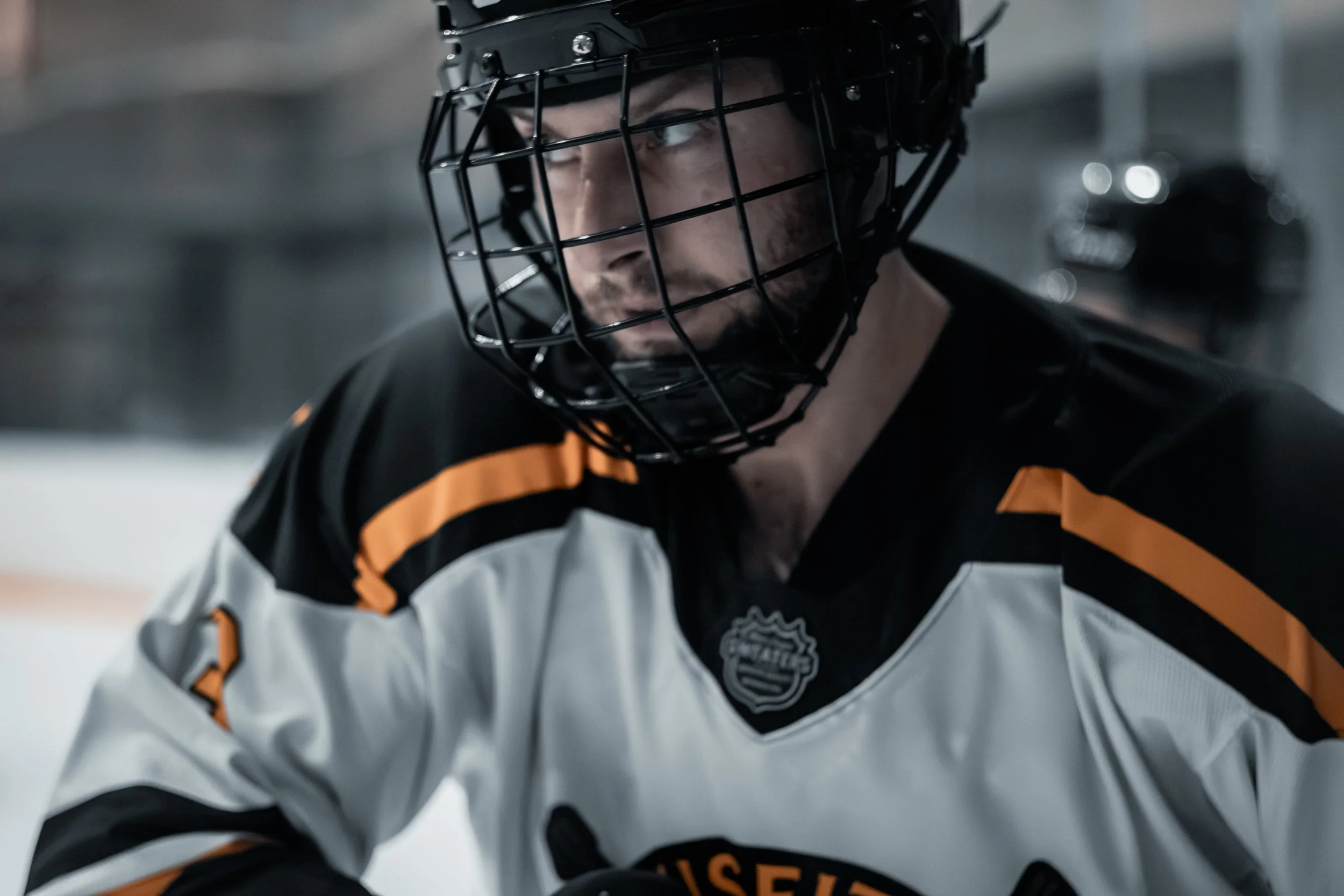 A young man in hockey gear, including a black helmet with a face cage, looking downward and to the side, inside a skating rink.