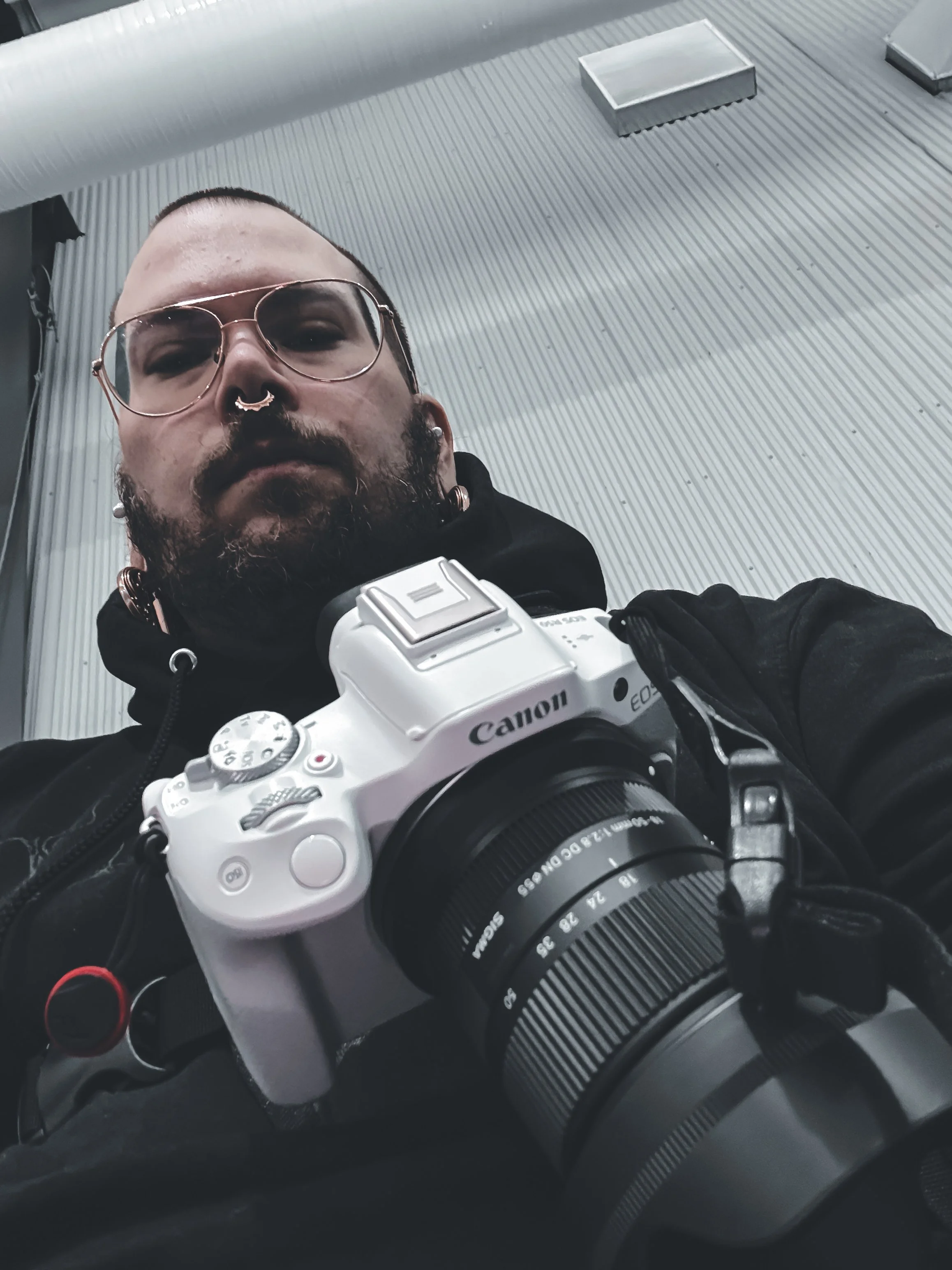 A person with sunglasses, facial piercings, and a beard holding a white Canon camera, taking a selfie in front of a metallic corrugated wall.