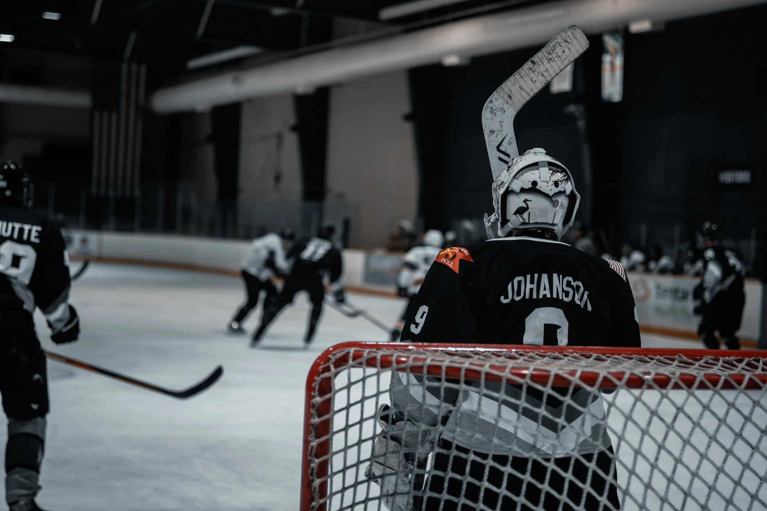 Hockey players on the ice during a game, viewed from behind a goal net with player Johanson. The rink is indoors with dark walls and overhead lights.