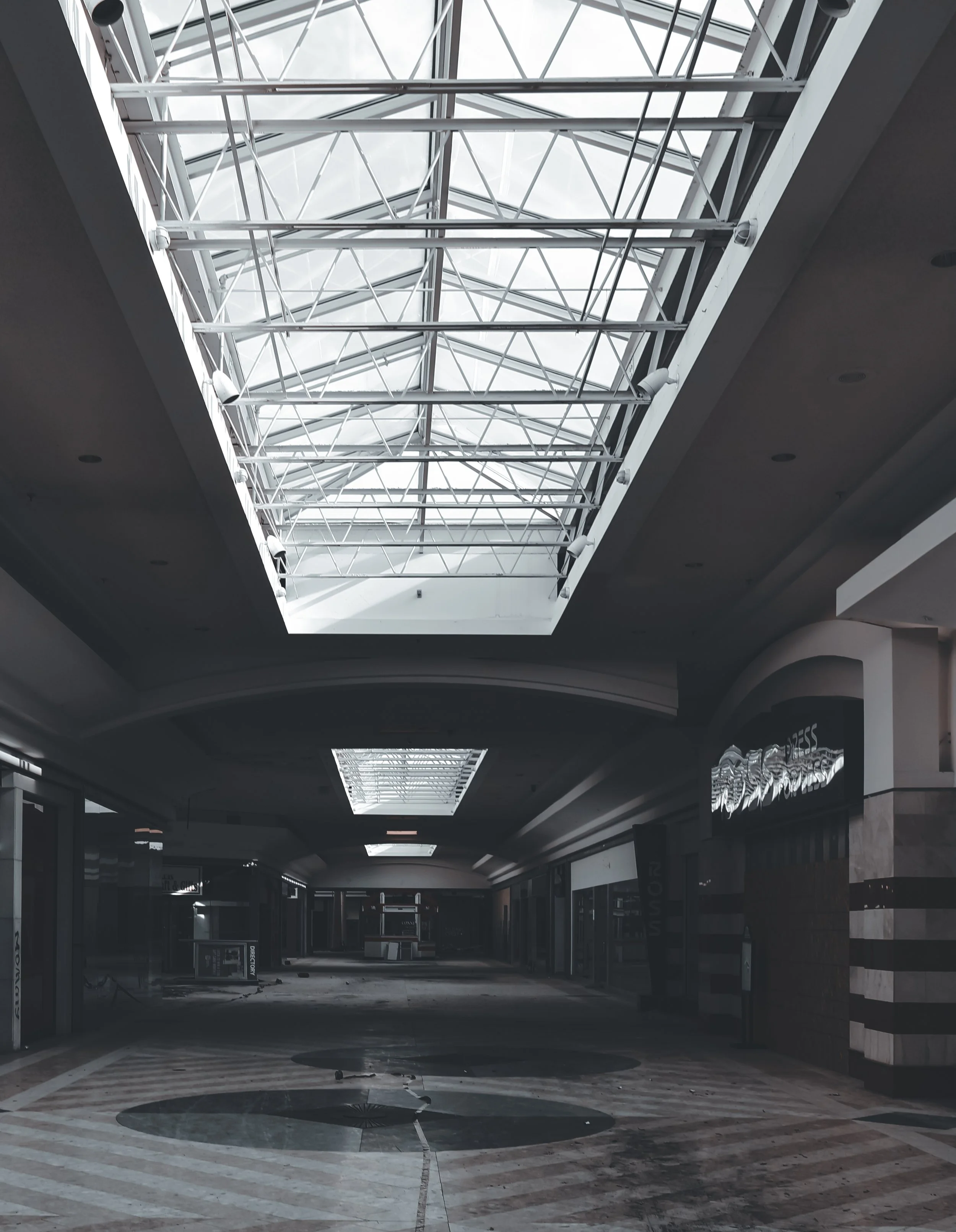 Interior view of a shopping mall with a large skylight ceiling and empty, dark storefronts.