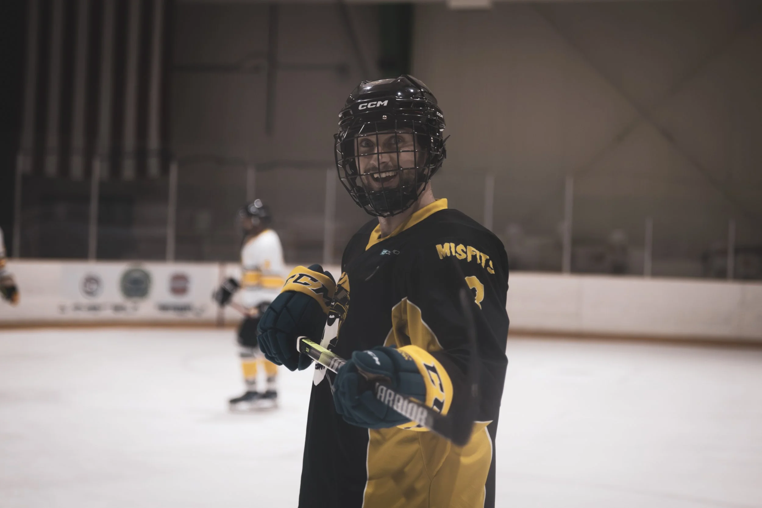 An ice hockey player wearing a black and yellow jersey and a black helmet on an ice rink, smiling and looking at the camera.