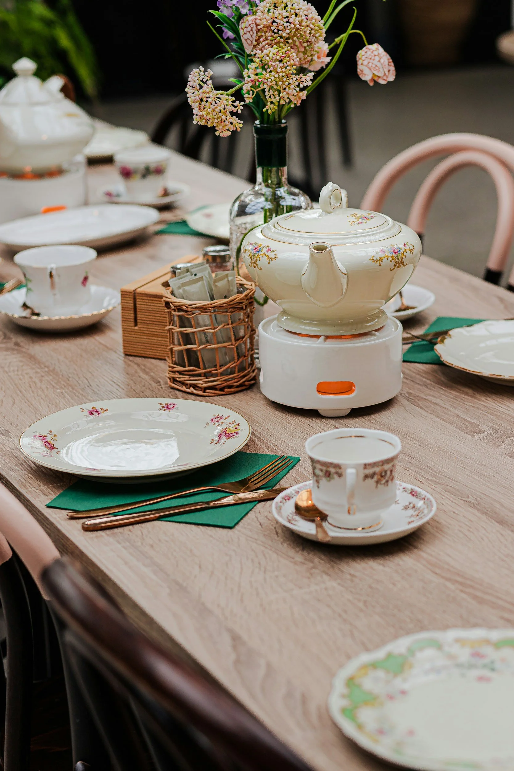Mismatched floral china, teacups, and a teapot arranged with green napkins on a softly styled table.