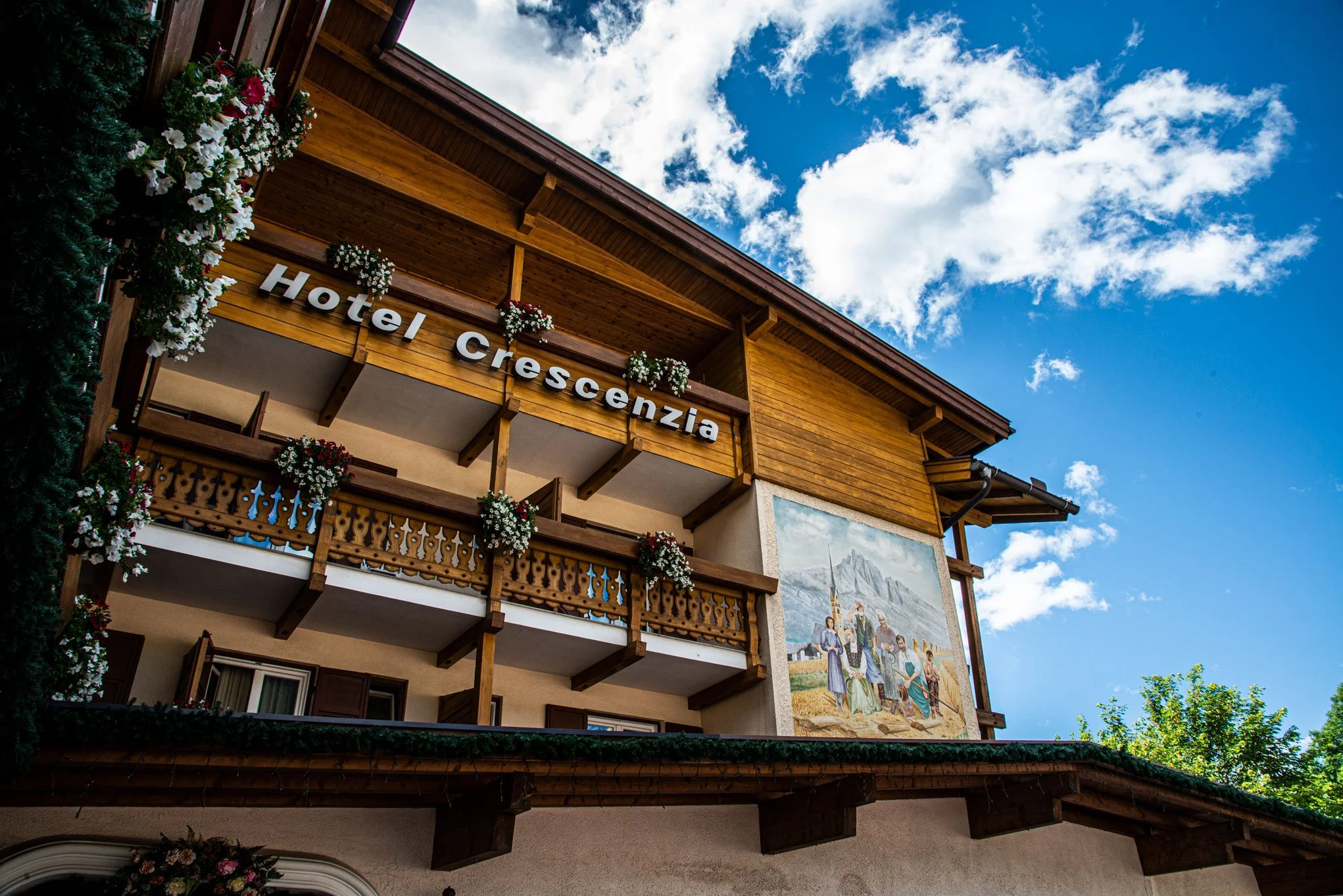 Facciata di un hotel in stile alpino in legno, con balconi decorati con fiori, sotto un cielo blu con nuvole, e un murale che raffigura scene tradizionali