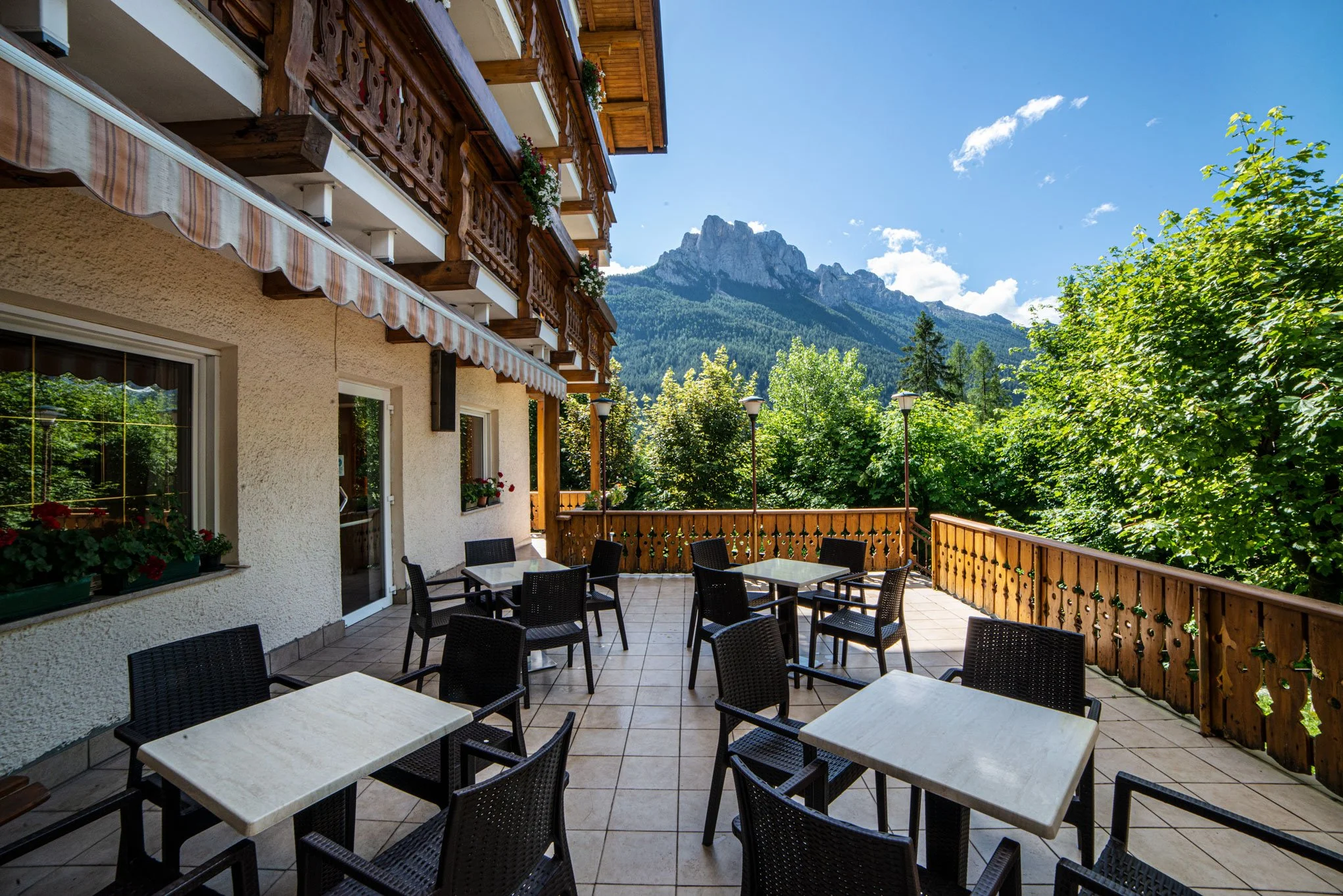 Terrazza di un albergo o ristorante con tavoli e sedie, ringhiere in legno, piante sui davanzali, e montagne sullo sfondo sotto un cielo sereno con alcune nuvole.