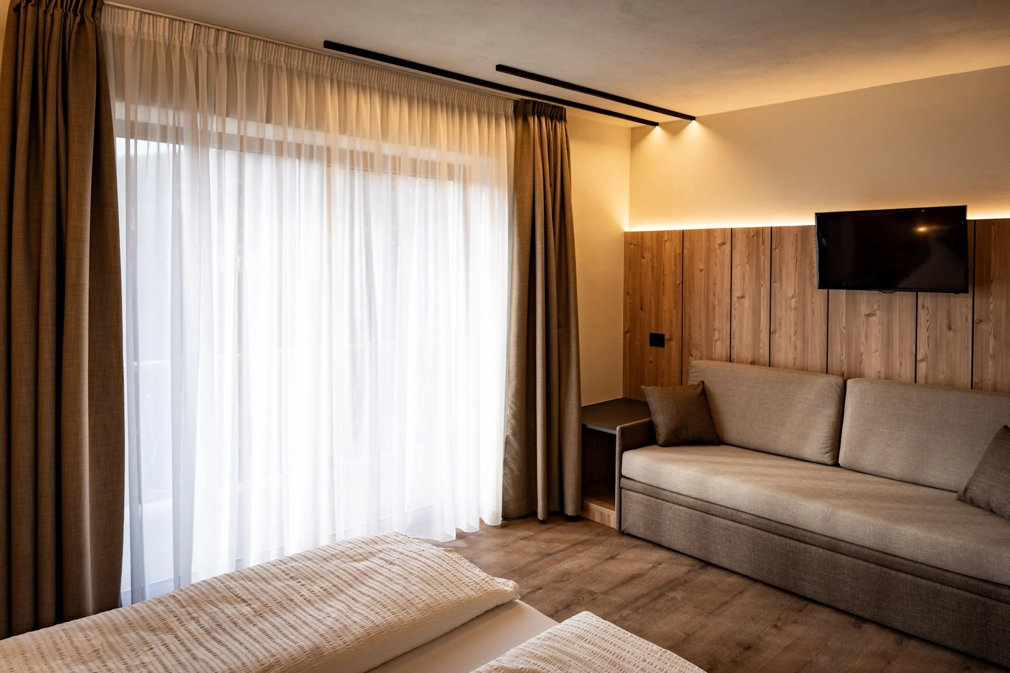 Interno di una stanza con tende bianche e marroni, pareti beige, pavimento in legno, divano beige e televisore montato sulla parete di legno.