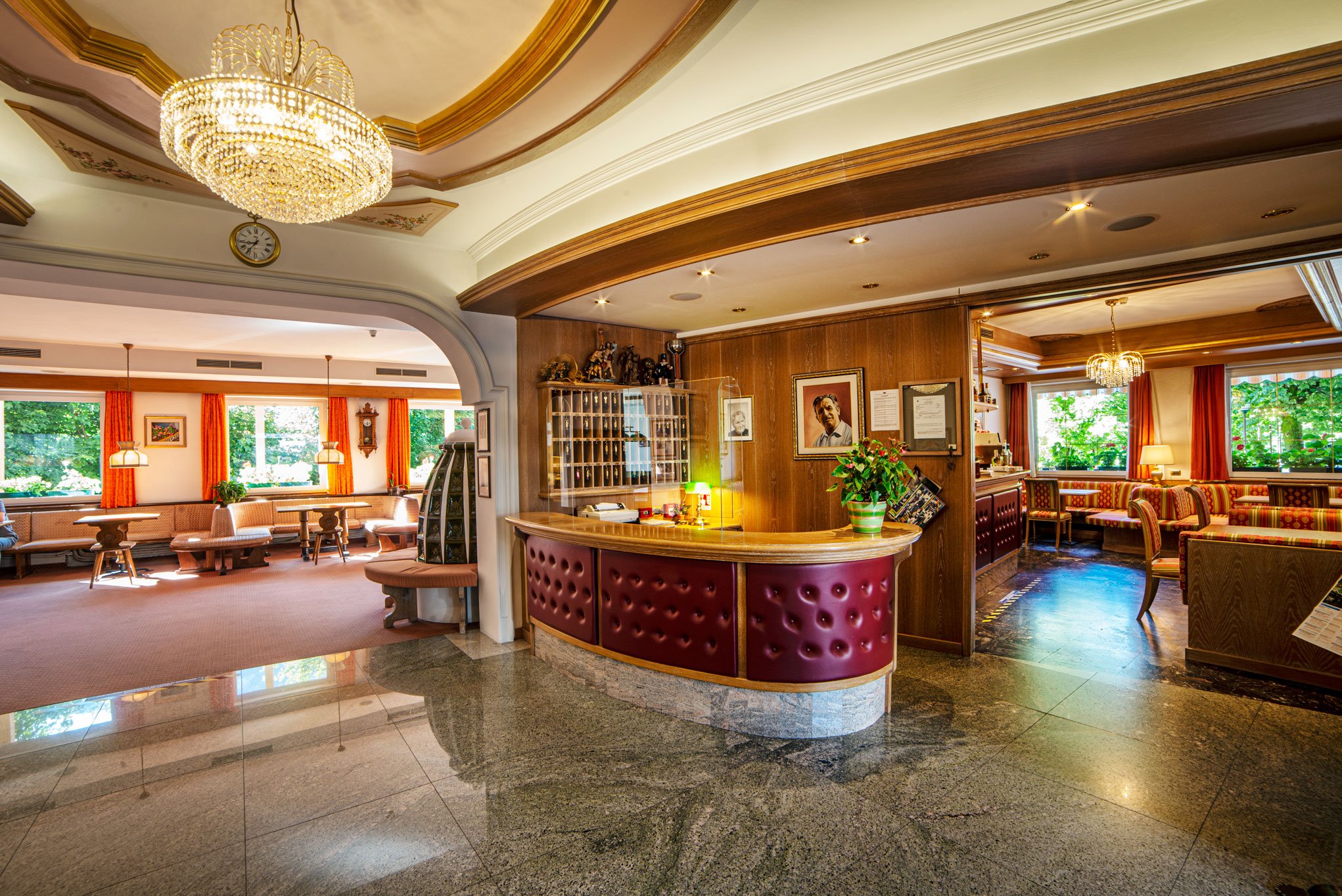 Interno di un bar o caffetteria con pareti in legno, un bancone curvo in pelle rossa, molte sedie e tavoli, quadri alle pareti, grandi finestre con tende arancioni, lampade e lampadari decorativi, una parete con fotografie e oggetti decorativi, e un 