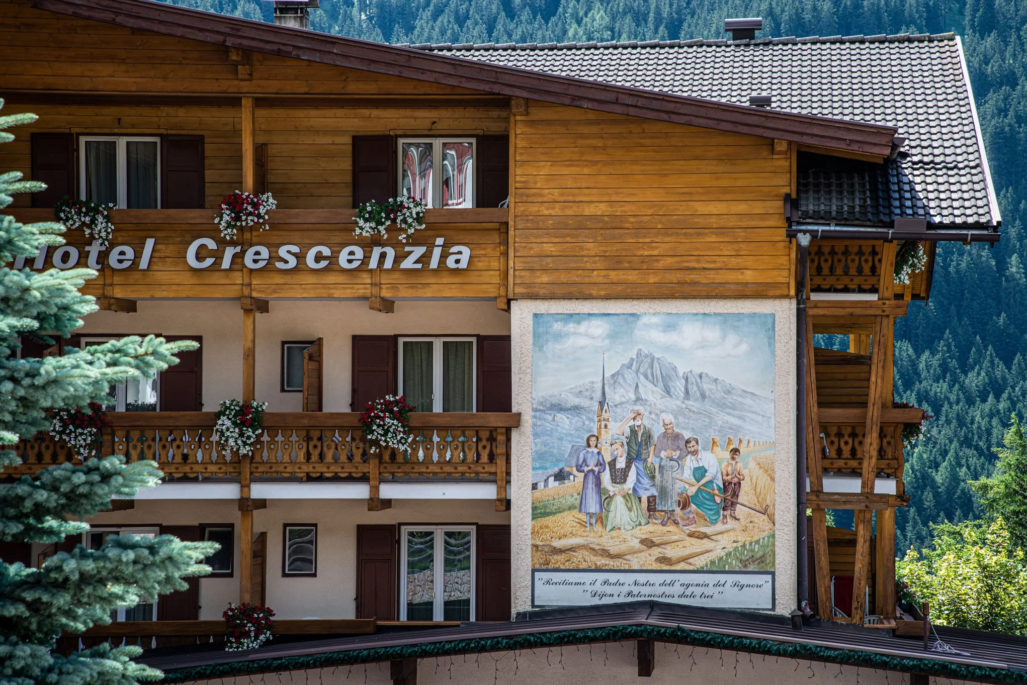 Edificio in stile montano in legno con balconi decorati con fiori. Sulla parete del palazzo c'è un grande dipinto che mostra persone in un campo con montagne sullo sfondo, e una scritta in italiano.