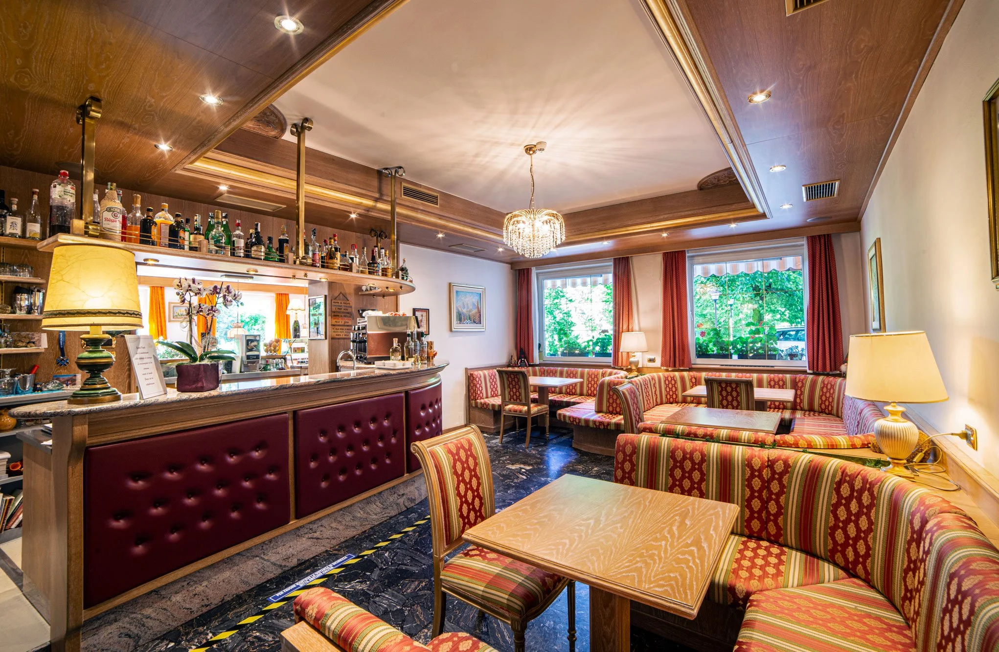 Interno di un bar o caffetteria con sedie e tavoli con rivestimenti a righe colorate, ben arredato con tende rosse, lampade e quadri alle pareti, e un bar con bottiglie di alcolici e bicchieri dietro il bancone.