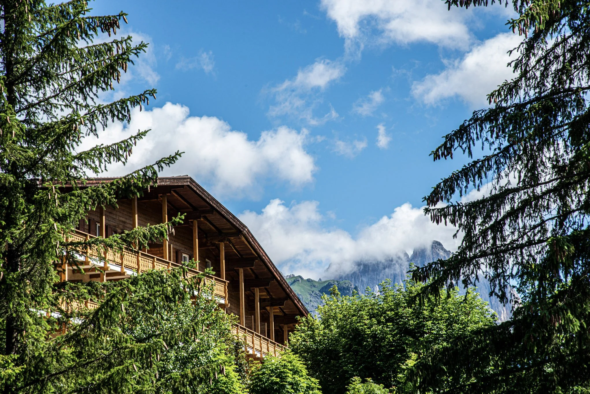 Casa di legno tra gli alberi in un paesaggio montano con cielo azzurro e nuvole.