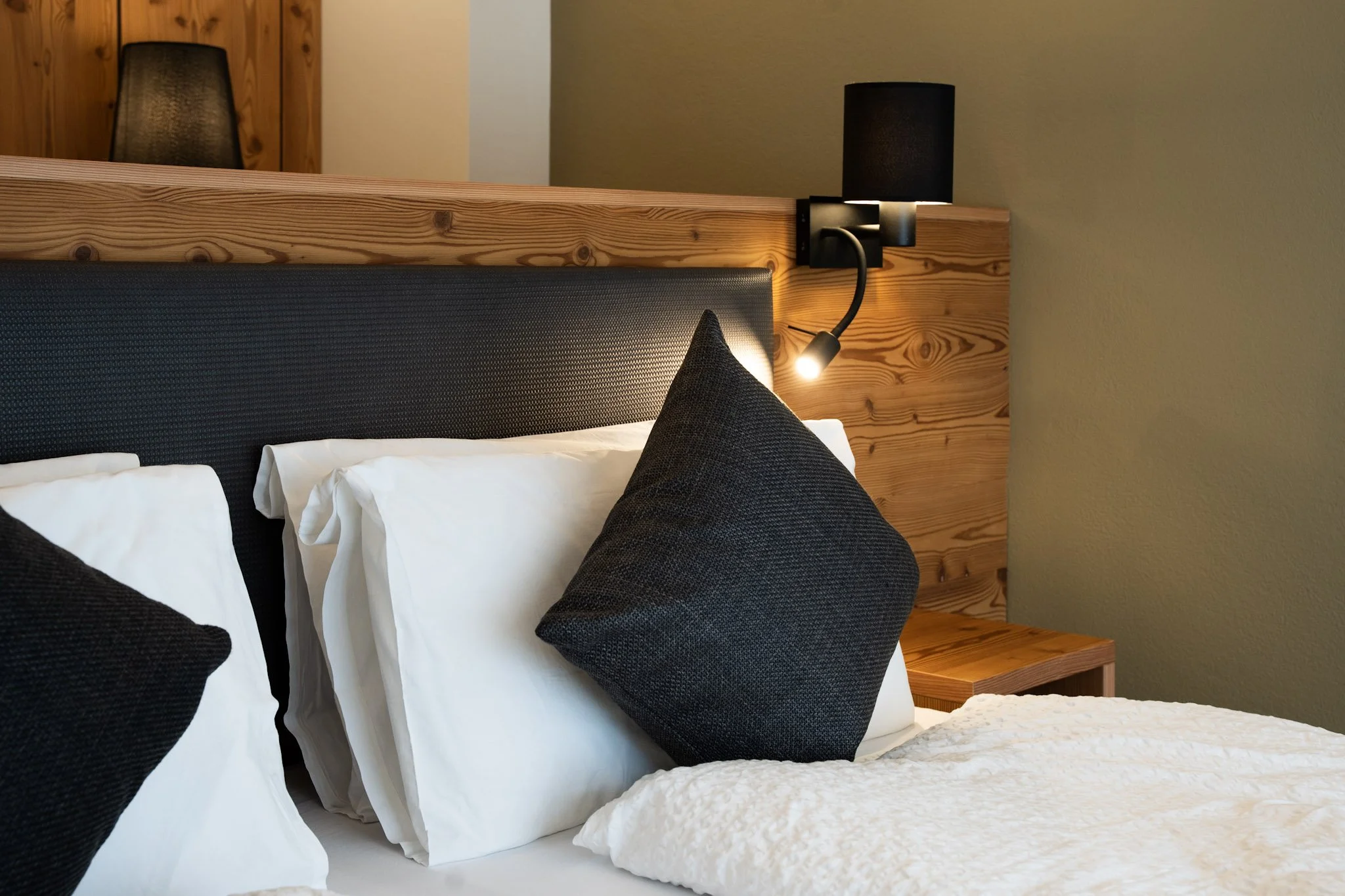 Letto con cuscini bianchi e copriletto bianco, testiera imbottita nera, lampada da parete nera accesa, parete di colore beige e testiera di legno chiaro in uno stile rustico.