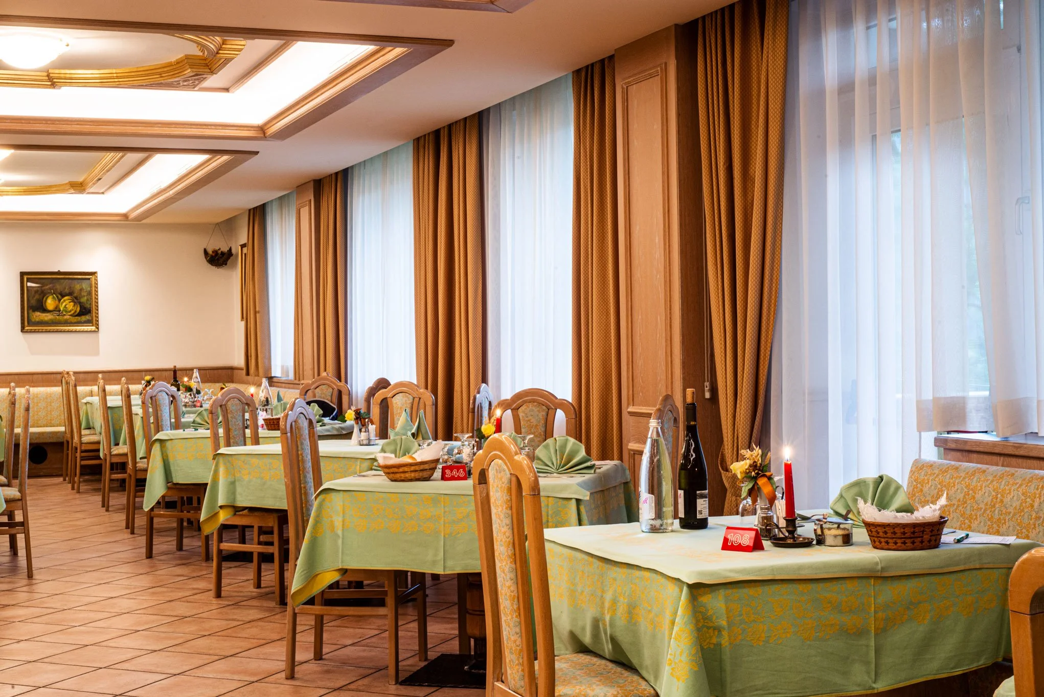 Interno di un ristorante elegante con tavoli apparecchiati con tovaglie verdi, piatti, bicchieri e decorazioni floreali. Le pareti sono decorate con quadri e ci sono grandi tende di tessuto che coprono le finestre, creando un'atmosfera accogliente.