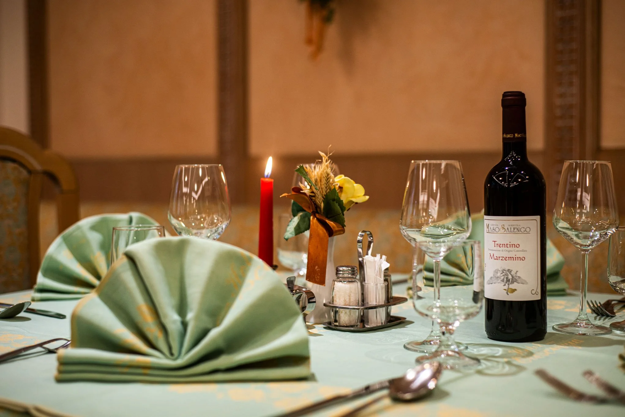Tavolo apparecchiato con bicchieri, posate, un centrotavola con fiori e candela rossa, bottiglia di vino, tovaglioli piegati, sale e pepe, in un ambiente elegante.