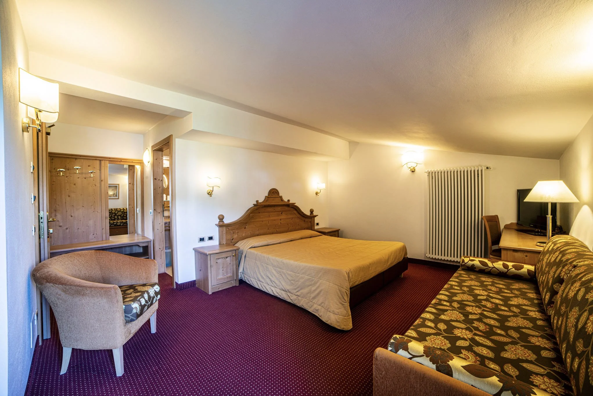 Camera di hotel con letto matrimoniale, armadio, televisione, sedie, lampade e rivestimenti in legno.