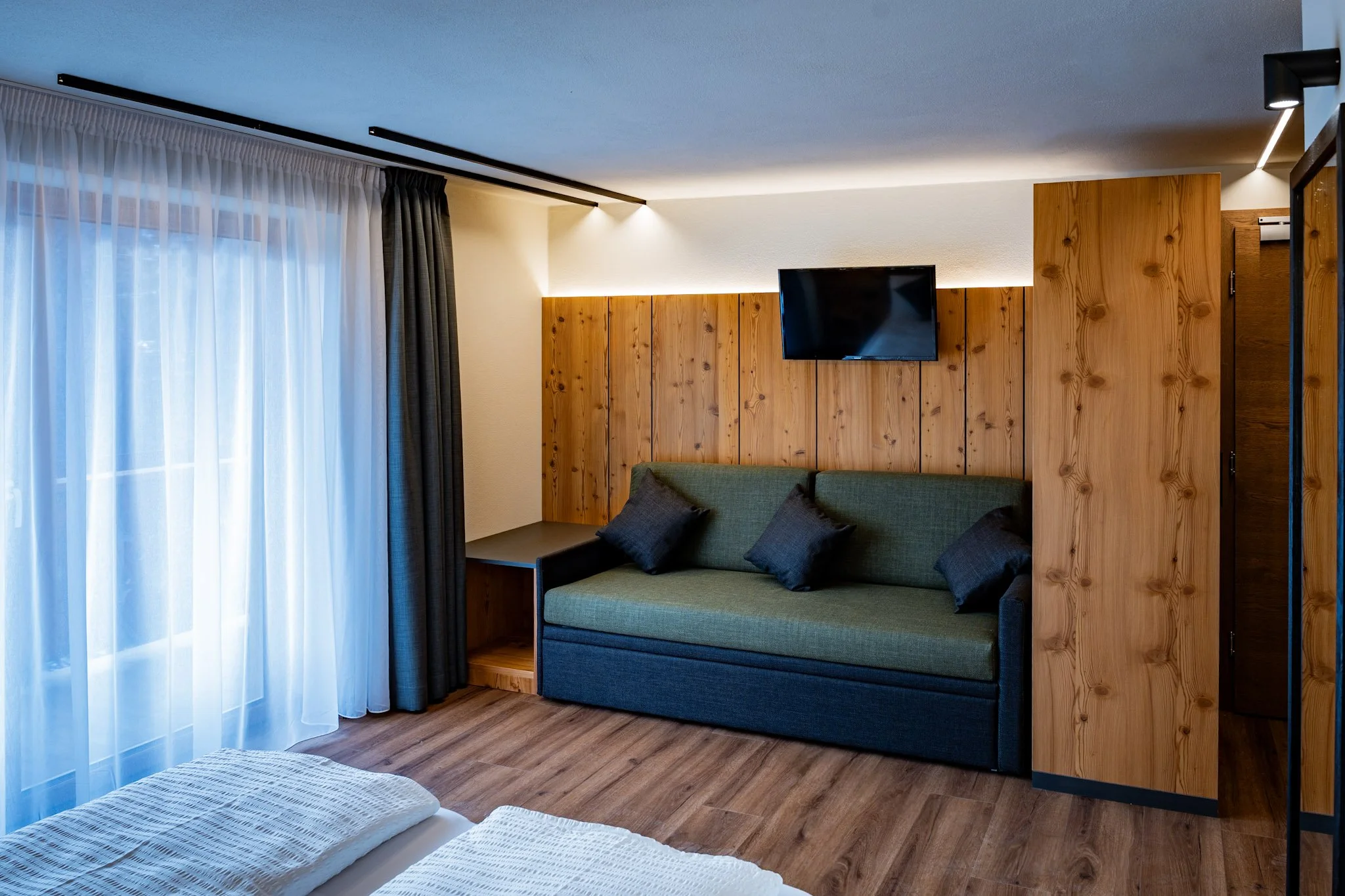Interno di una stanza con parete in legno, divano verde con cuscini neri, tende di tessuto bianco e scuro, TV a parete e pavimento in legno.