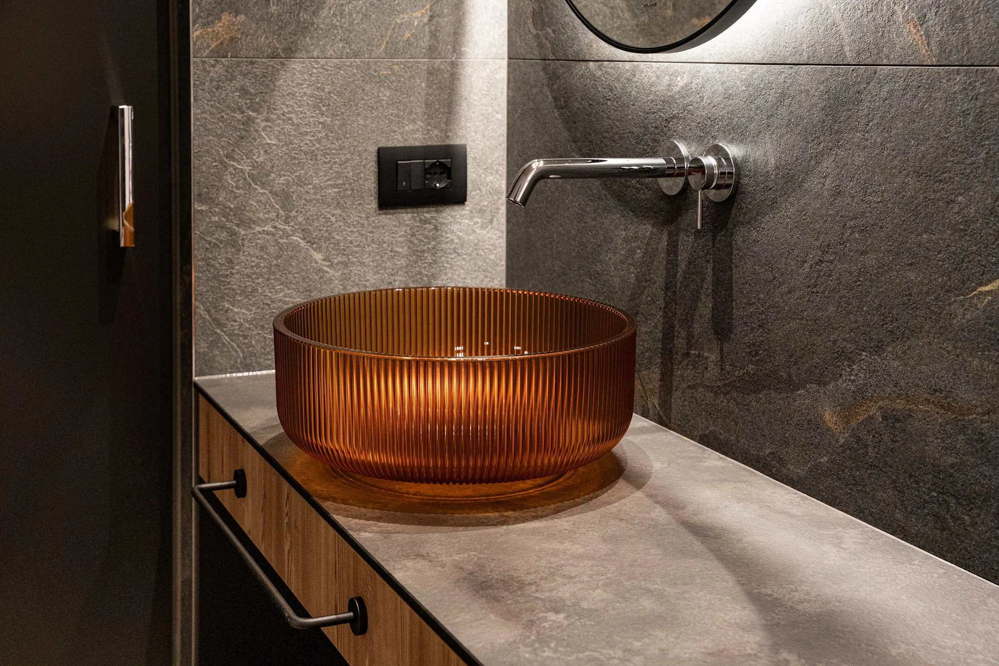 Un lavandino rotondo in ceramica color rame con superficie a righe verticali, posizionato su un mobile di legno chiaro, con pareti in pietra grigia e un interruttore elettrico nero sopra il lavandino.