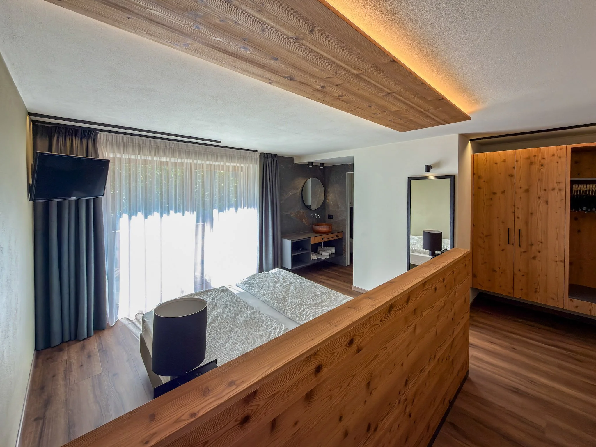 Interior di una camera con pareti bianche, pavimento in legno e un soffitto decorato con pannelli di legno e illuminazione a incasso. Una grande finestra con tende leggere permette l'ingresso di luce naturale.