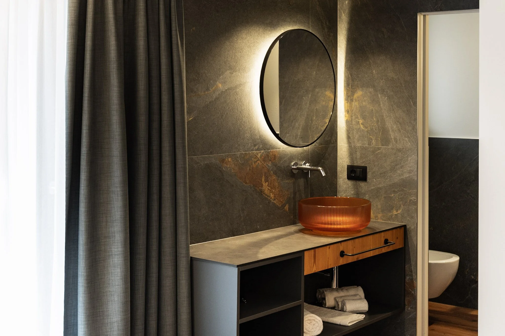 Specchio rotondo con retroilluminazione su muro di pietra, lavabo in ceramica arancione con pila in legno e armadietto in legno, tende grigie e porta aperta che porta a un bagno.