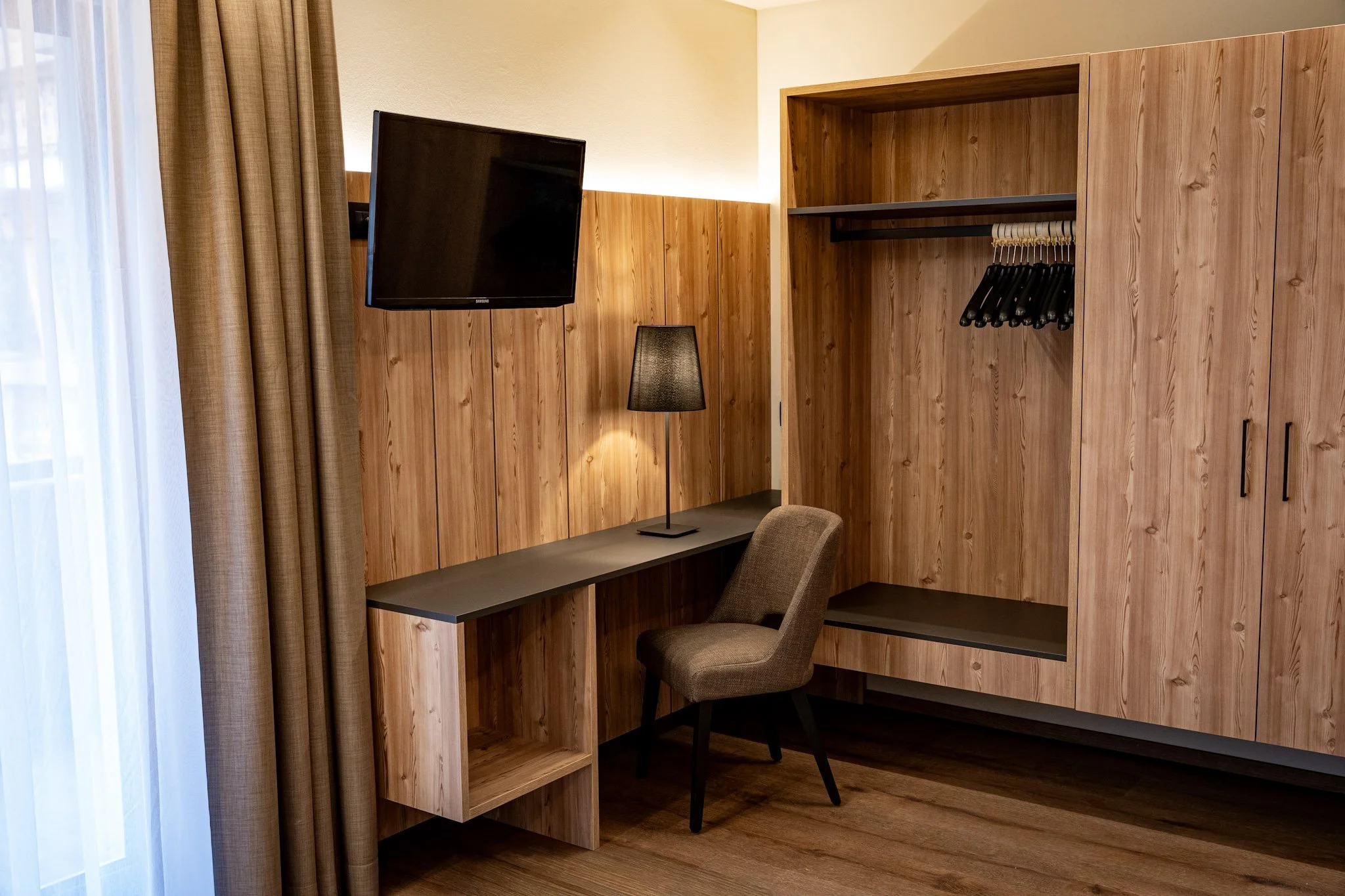 Camera di hotel con pareti di legno, scrivania, sedia, lampada da terra e armadio aperto con appendiabiti. Tenda davanti a una finestra.