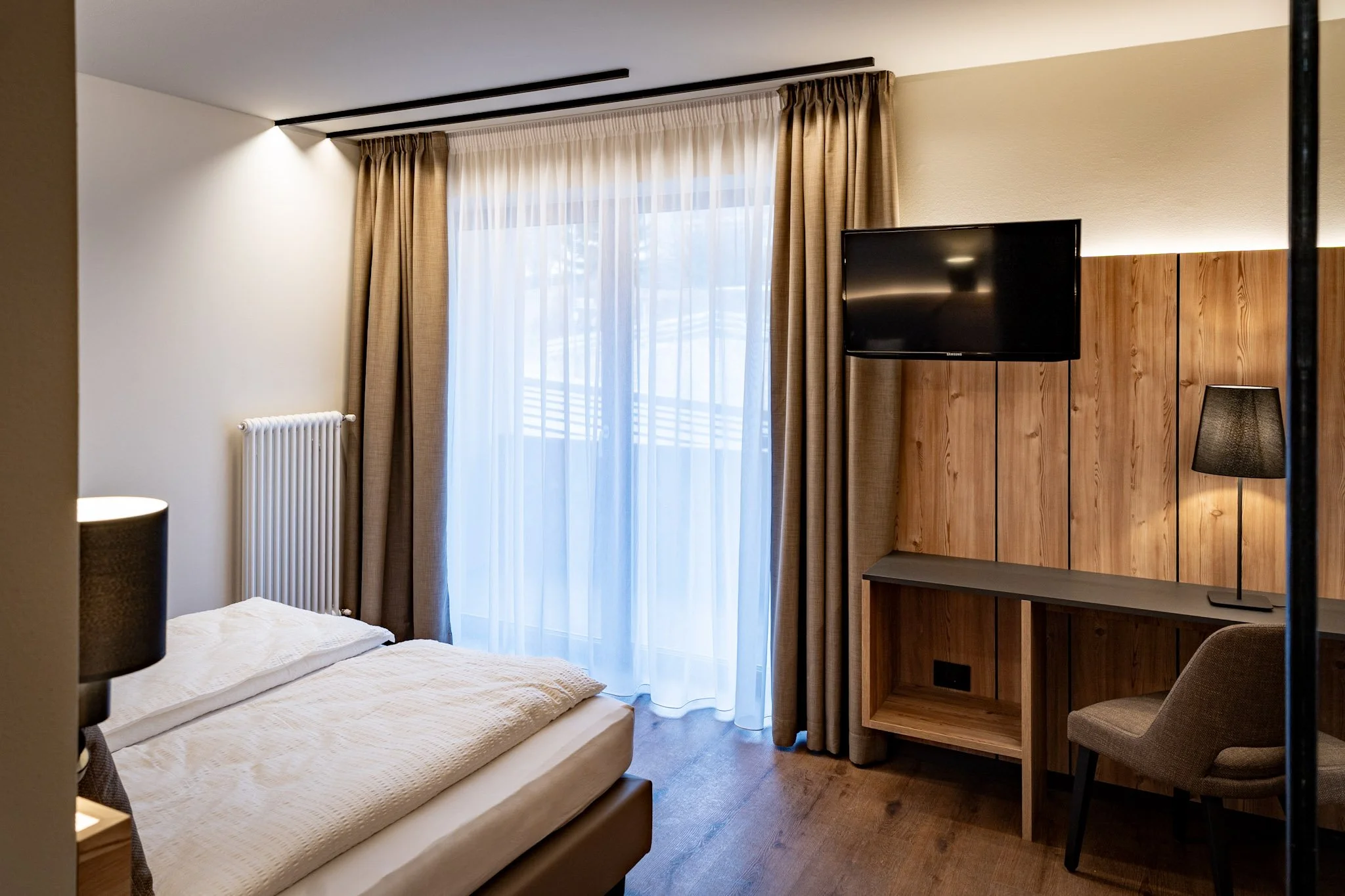 Camera da letto con un letto con materasso coperto da una coperta beige, una lampada da tavolo accesa, tende beige e un televisore nero appeso al muro di legno.