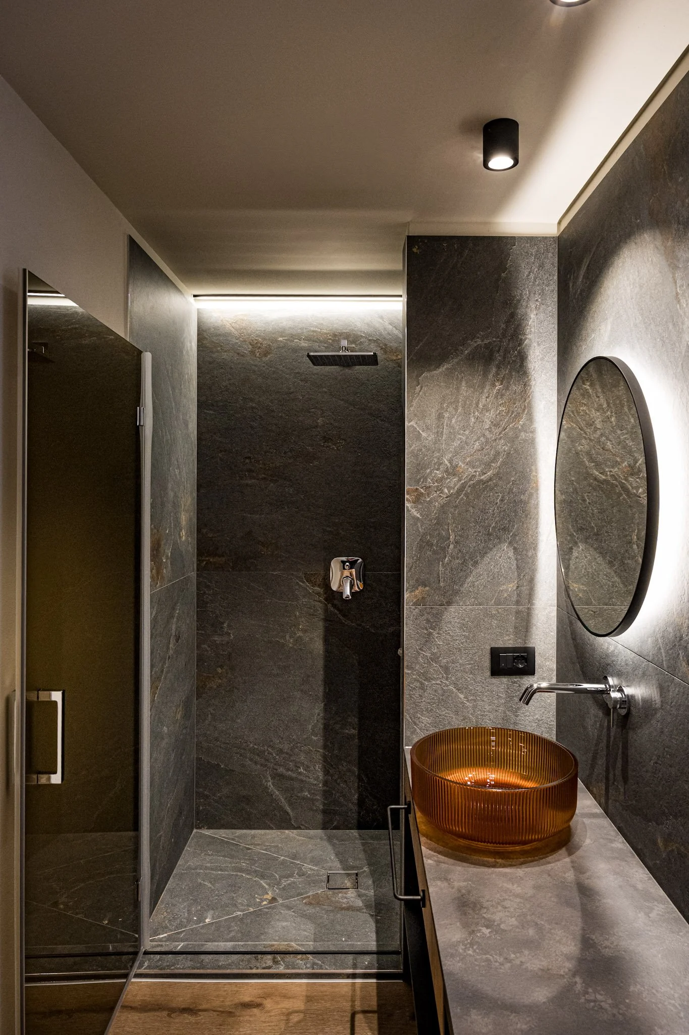 Interno di un bagno moderno con pareti e pavimenti in pietra scura, grande specchio rotondo e vaso arancione in vetro trasparente, illuminazione soffusa.