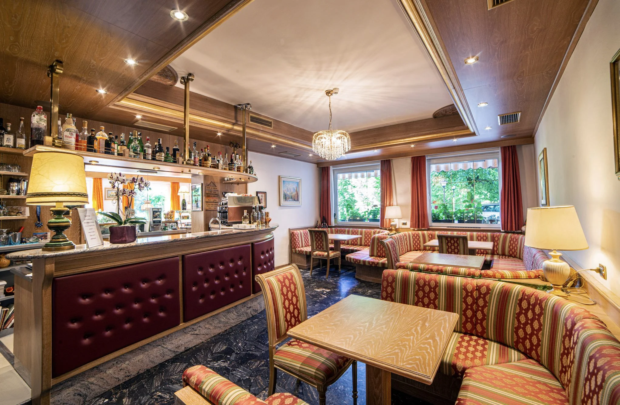 Interno di un ristorante con pareti rivestite in legno, tavoli con sedie a righe colorate e un bar con bottiglie di liquor e un grande lampadario al centro del soffitto.