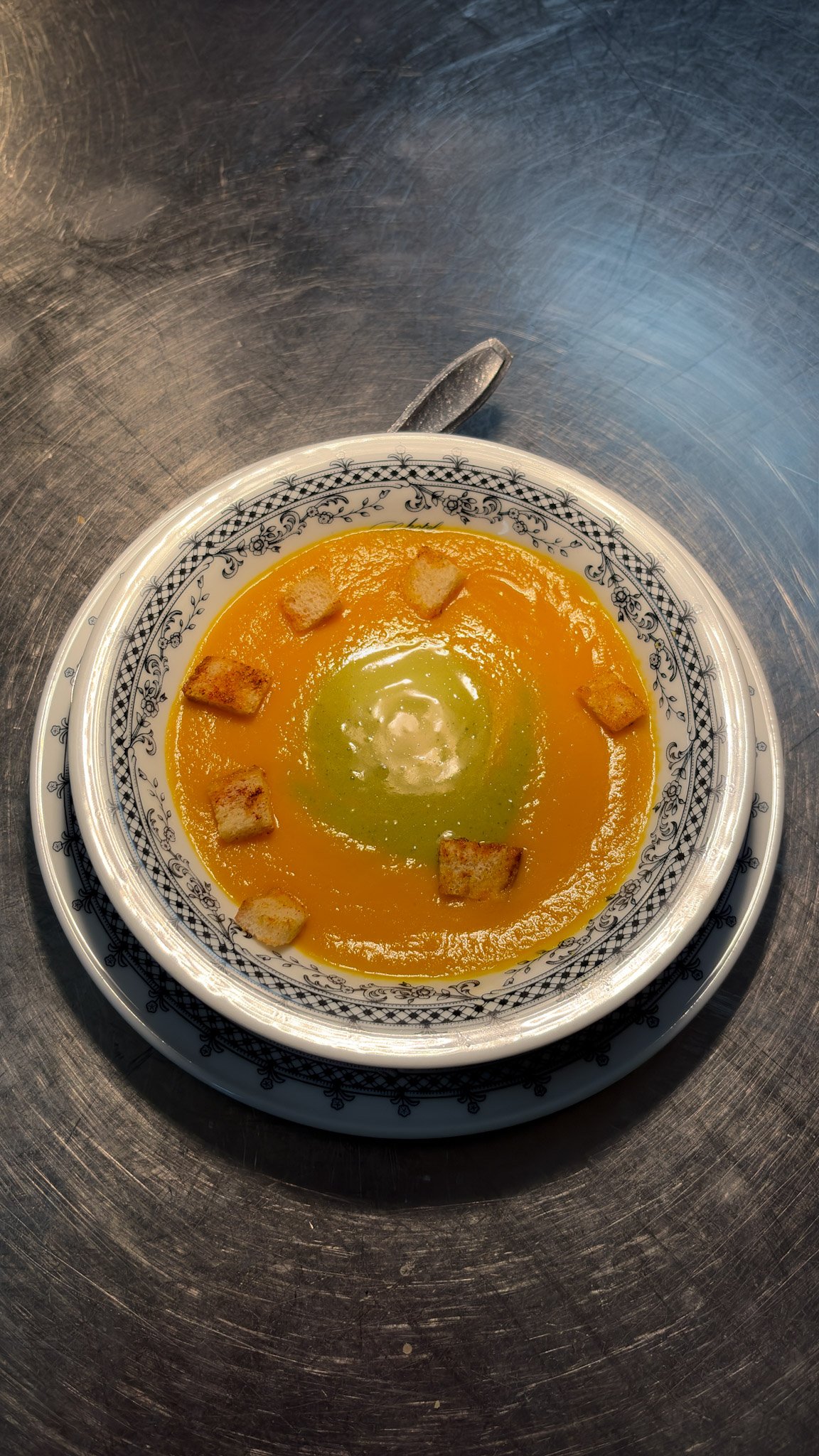 Zuppa di carote e zucca con crostini, servita in una ciotola decorativa con bordo nero e bianco su un piatto. Sfondo metallico.