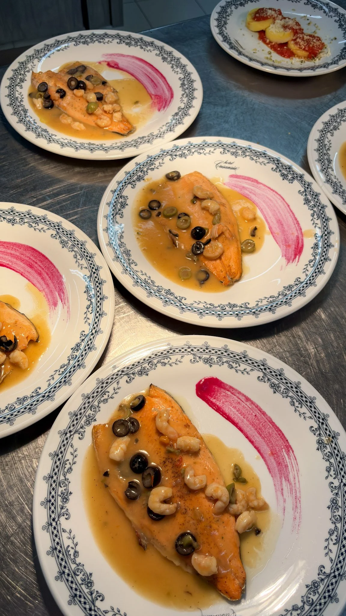 Filetto di salmone con salsa, olive nere e gamberi, servito su piatti bianchi decorati con motivo blu.