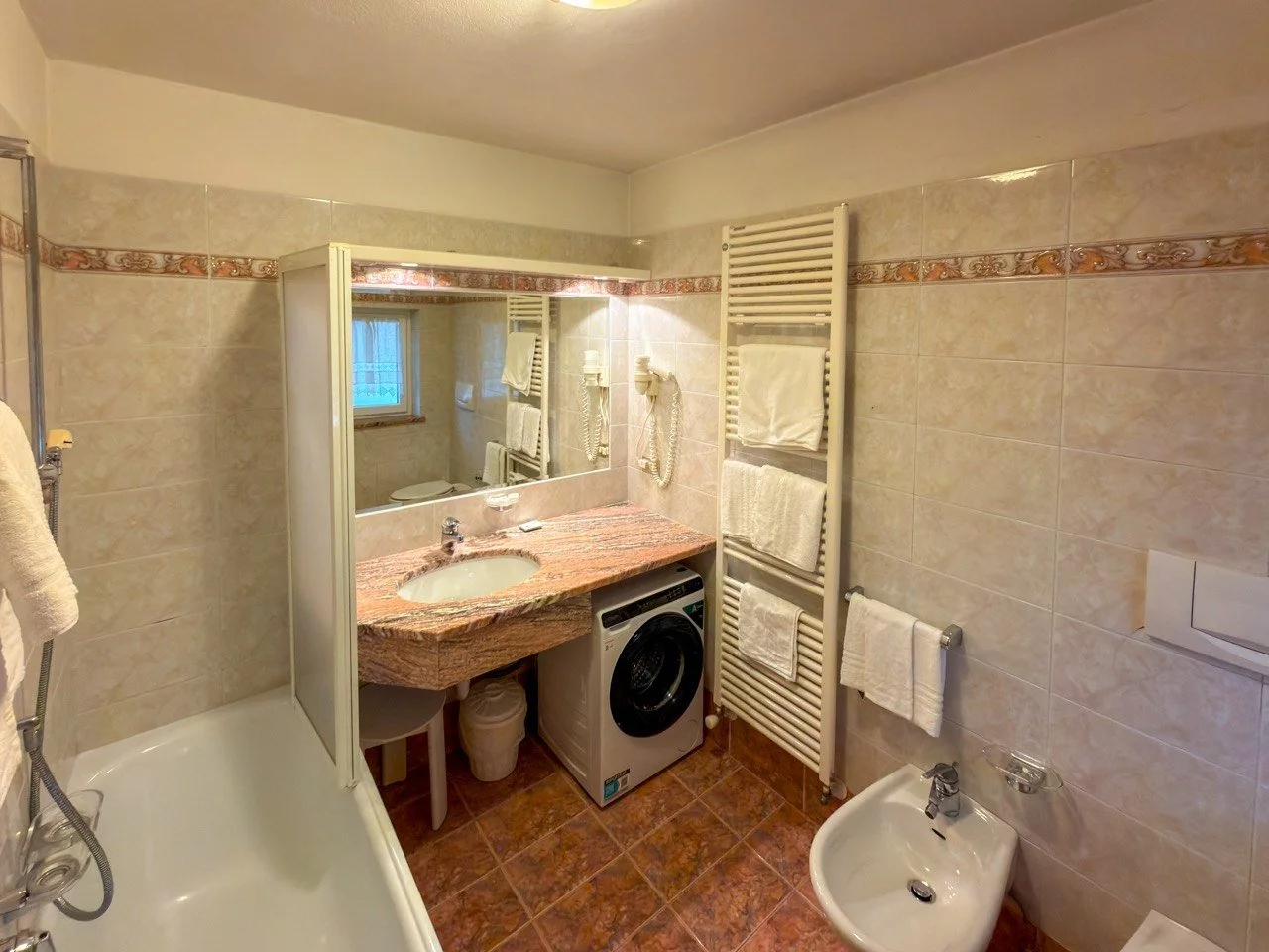 Bagno con lavabo in marmo, lavatrice, bidet, radiatore portasciugamani con asciugamani, specchio e finestra.