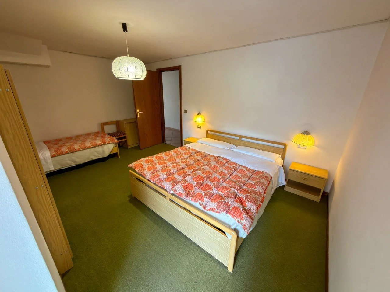 Camera con letto matrimoniale, lampade accese, armadio, letto singolo e porta aperta, arredamento semplice e contrasto di colori caldi.