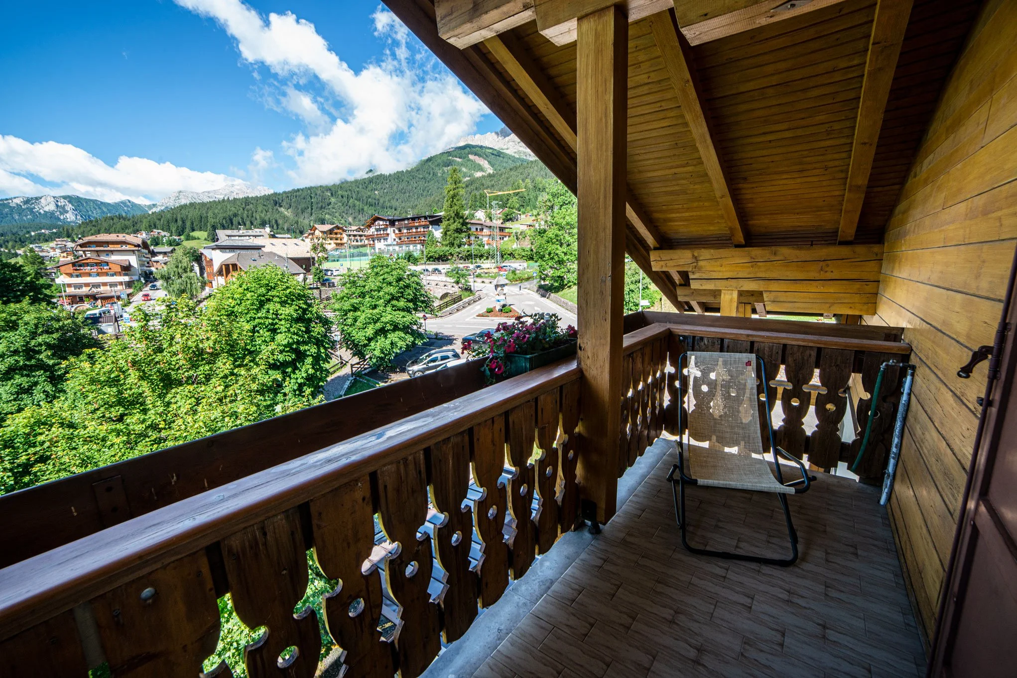 Vista da un balcone di una zona montuosa con case e alberi verdi, cielo blu con nuvole, e montagne sullo sfondo.