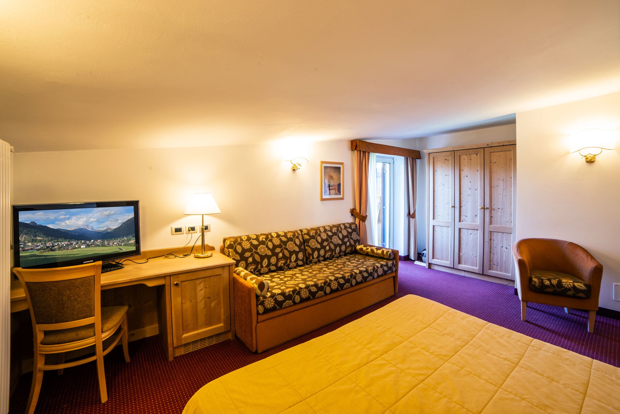 Interno di una stanza di hotel con un televisore, un divano, un armadio in legno e una porta finestra con tende. La stanza ha luci soffuse e uno stile caldo.