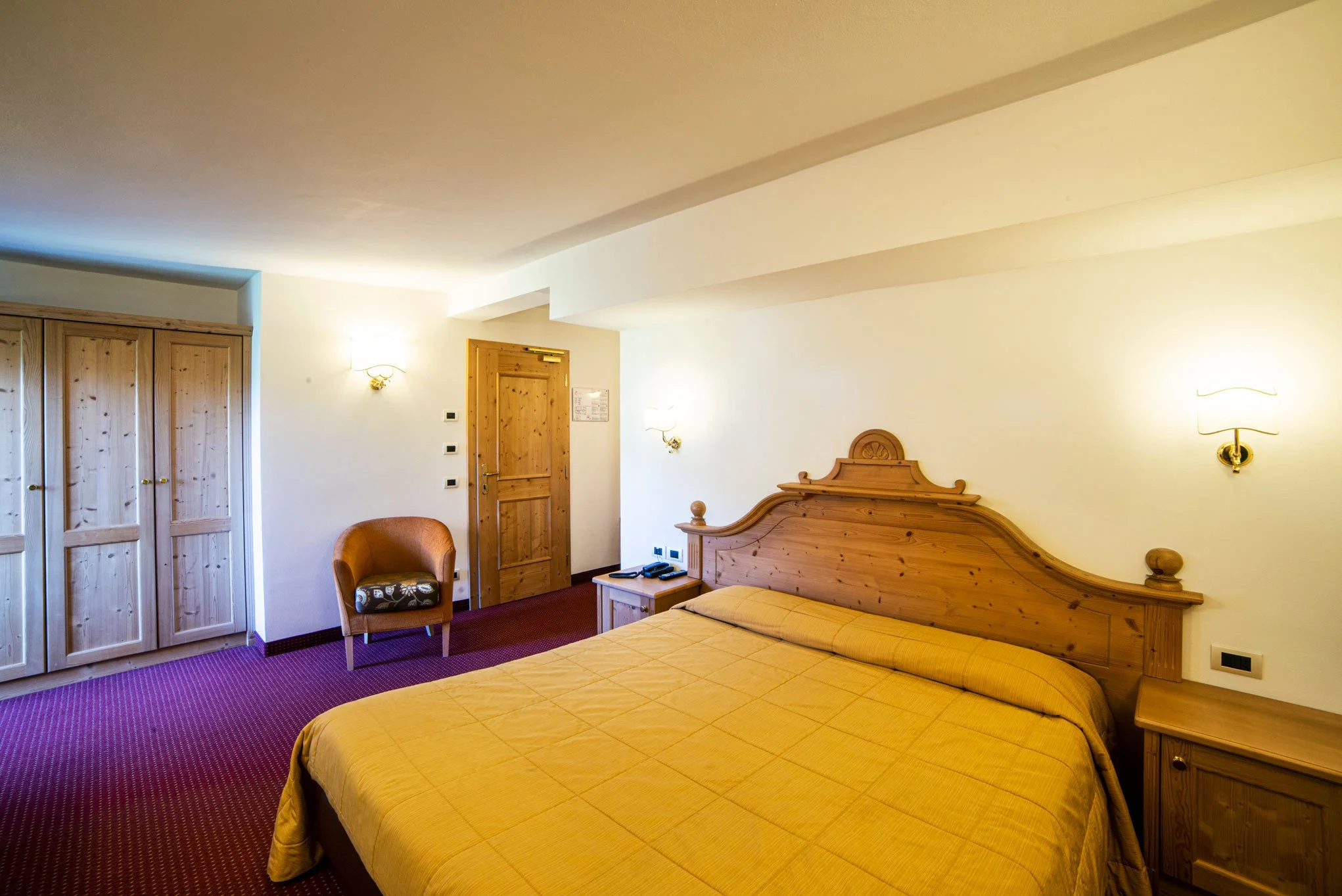 Camera da letto con letto matrimoniale in legno, armadio in legno, poltrona e pareti bianche con appliques luminose. Tappeto rosso e parete con porta di legno.