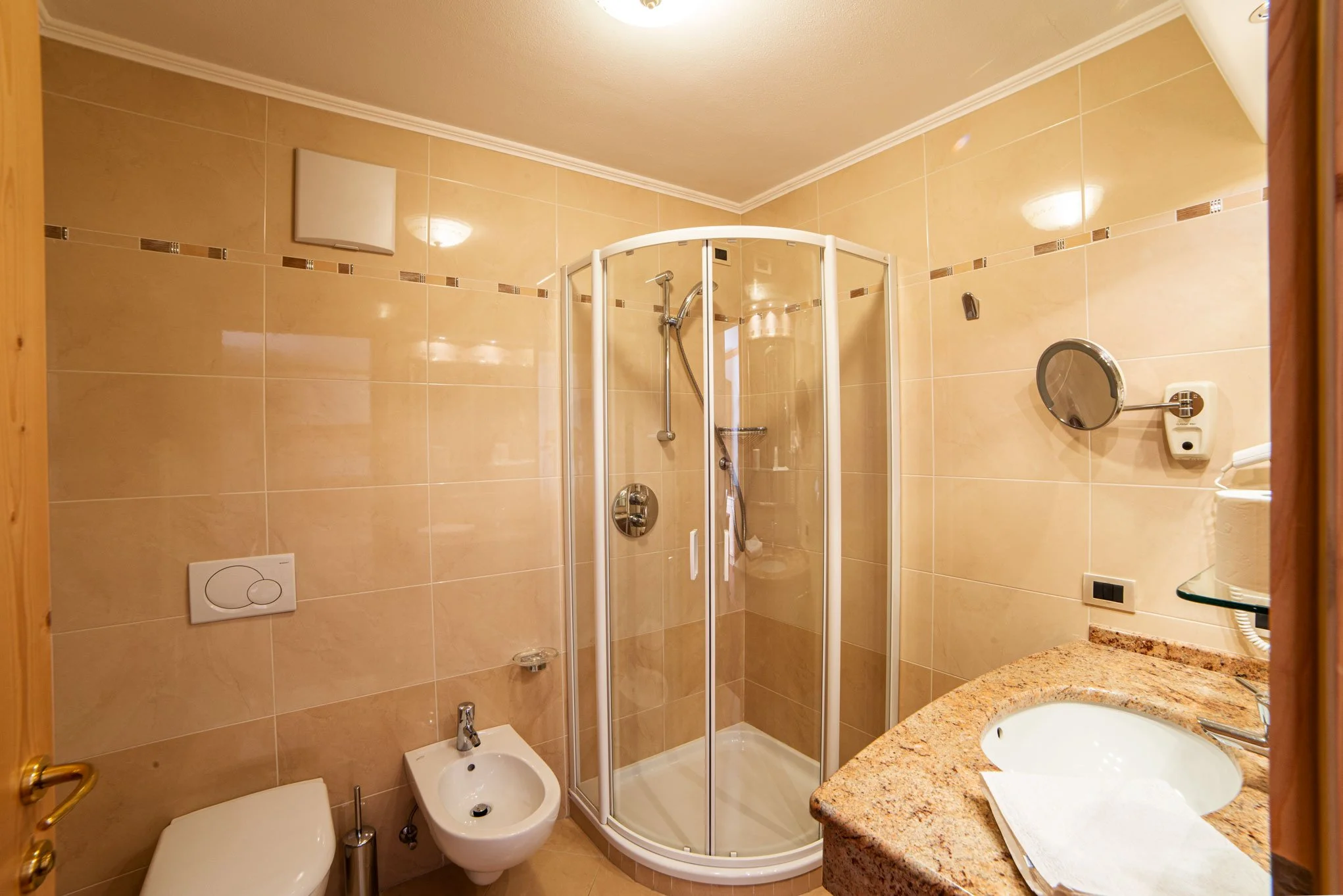 Bagno con pareti rivestite in piastrelle beige, doccia con porta a soffietto, lavabo con piano in granito, specchio rotondo e accessori per il bagno.