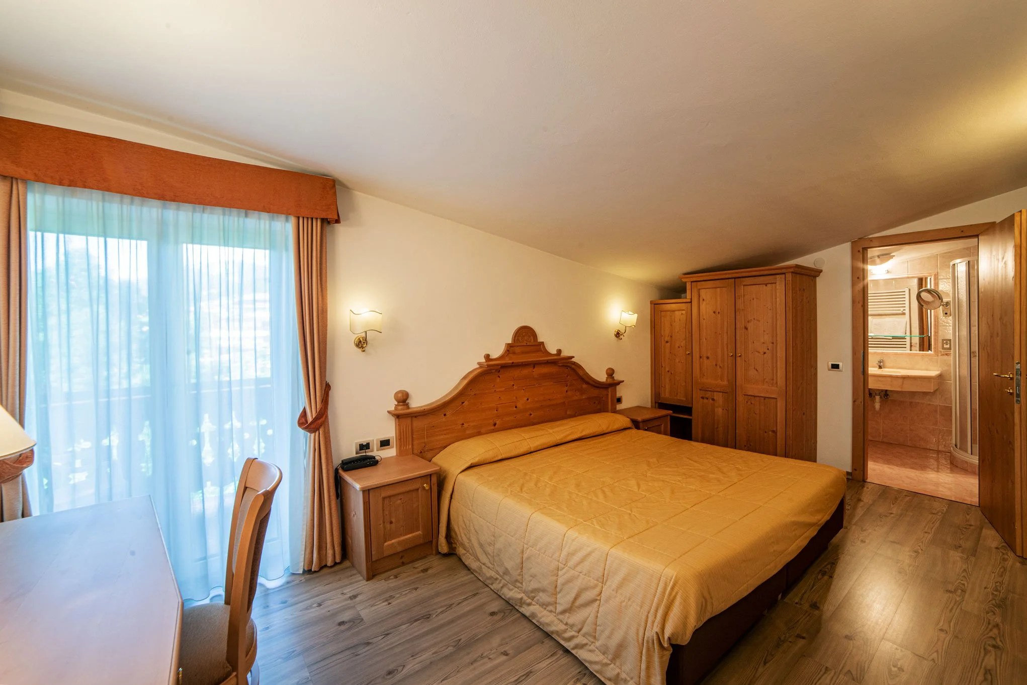 Camera di una stanza con letto matrimoniale, armadio in legno, e pareti illuminate da lampade. Una finestra con tende di tessuto leggero lascia entrare la luce naturale. Un bagno si intravede attraverso una porta aperta.