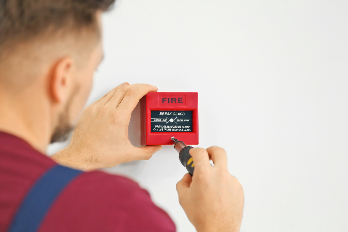 A person installing or repairing a fire alarm connected to a fire alarm box on a white wall.