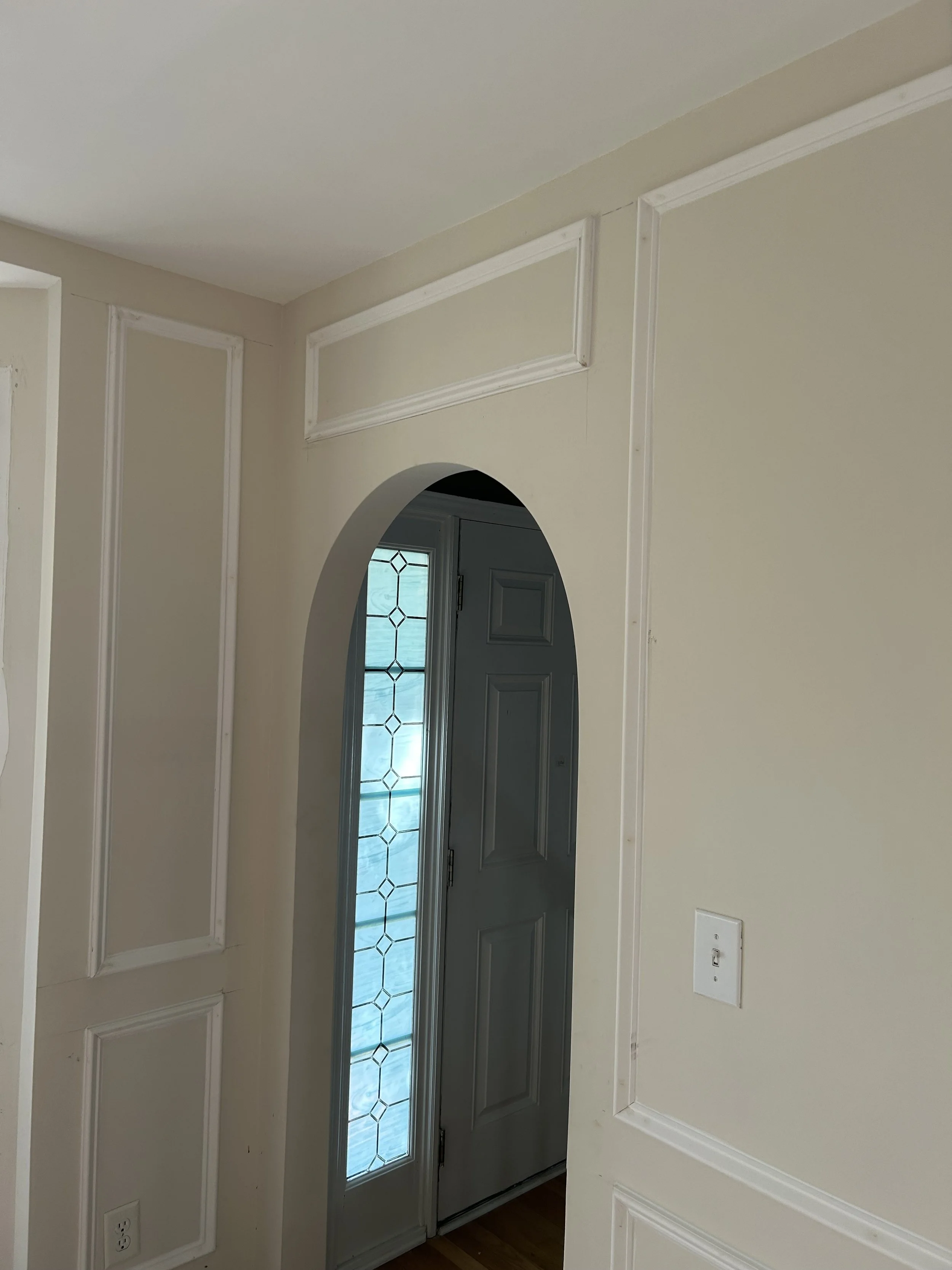 Interior view of an entryway with a front door featuring decorative glass panels, framed by cream-colored walls with white molding, and an arched doorway leading inside.