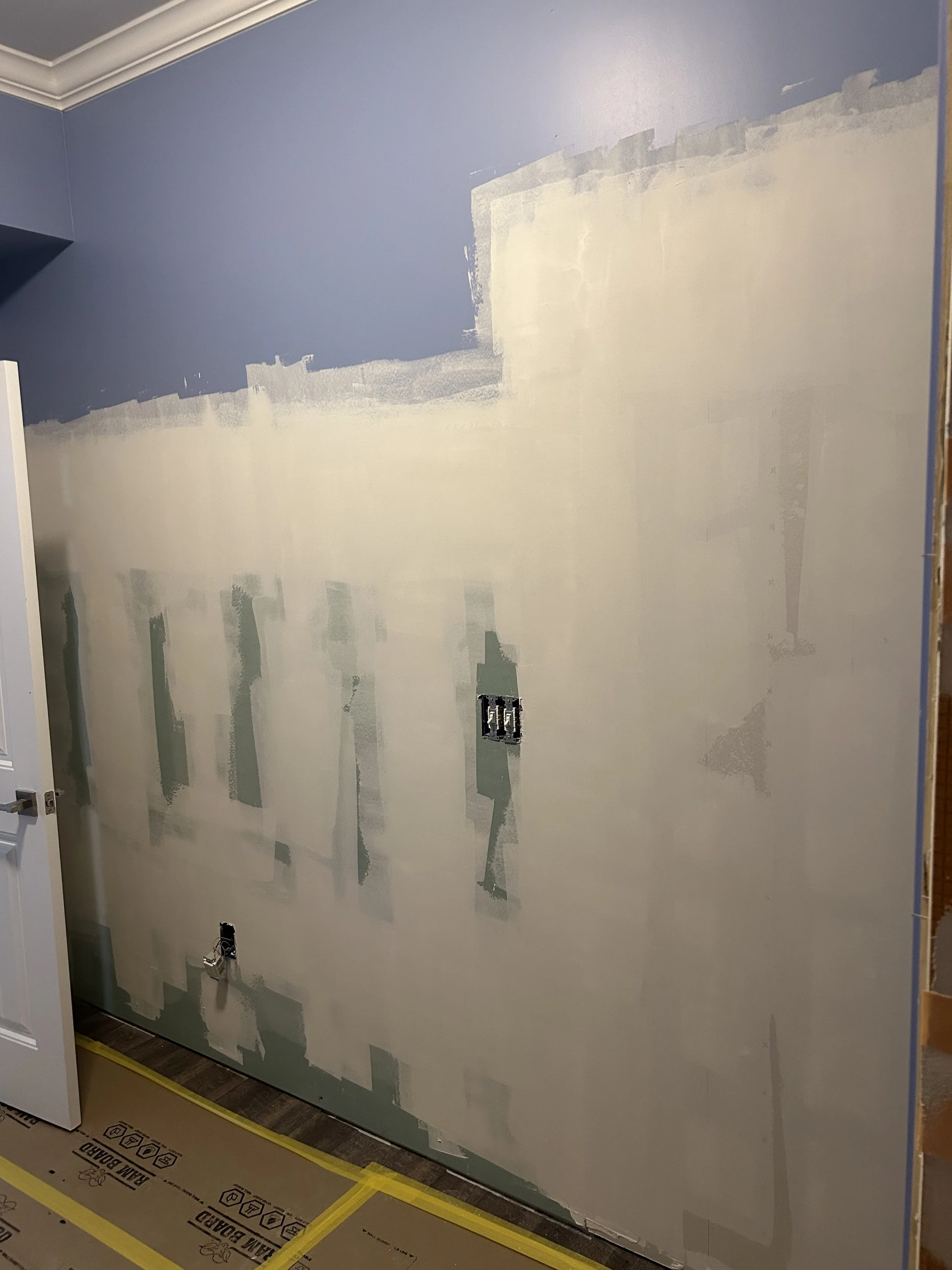 Interior wall undergoing renovation with drywall partially installed and taped, electrical outlets not yet installed, and floor covered with protective sheets.