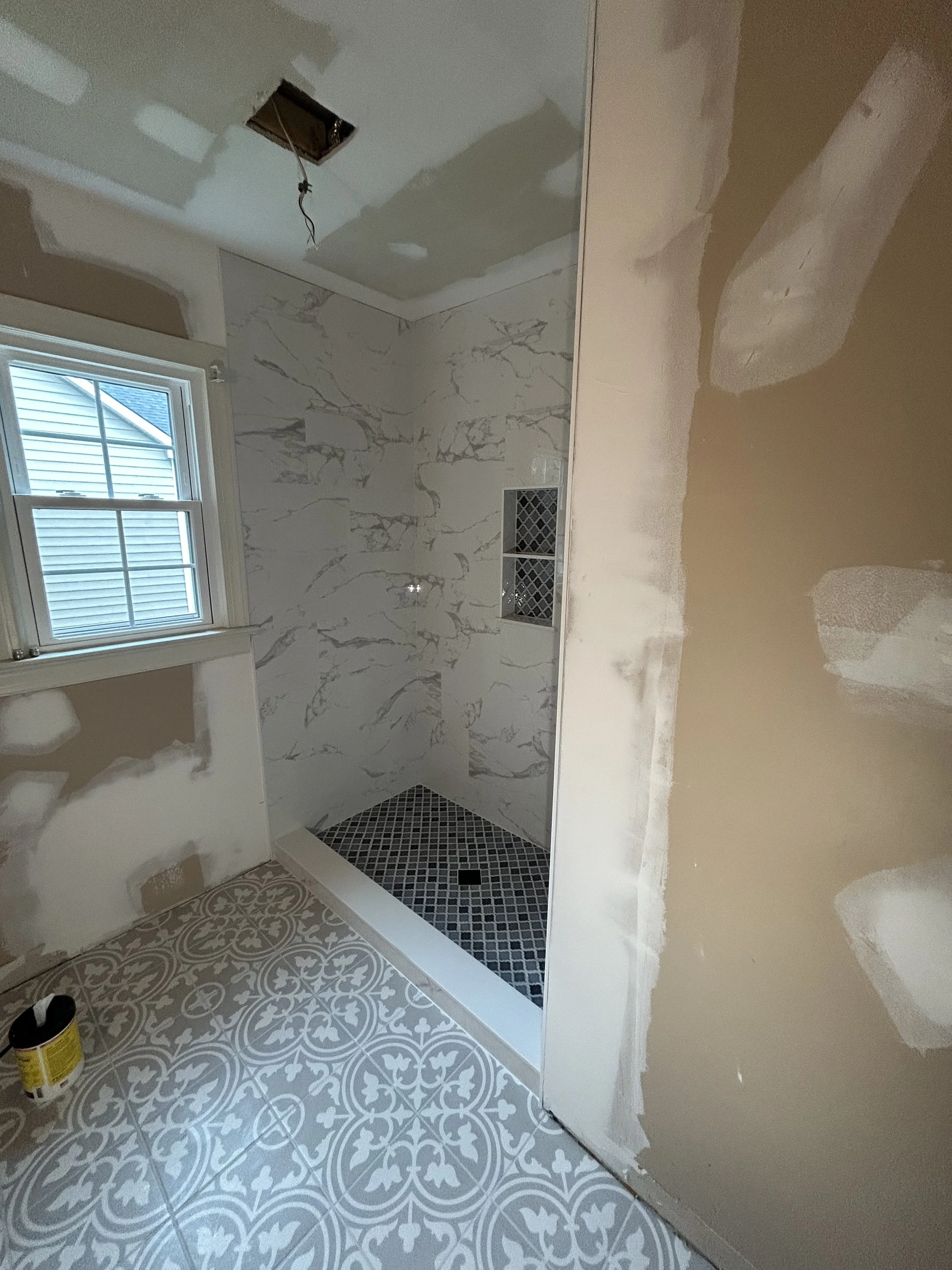 A bathroom under renovation with partially finished walls, a window, decorative patterned floor tiles, and an unfinished ceiling with exposed wiring.