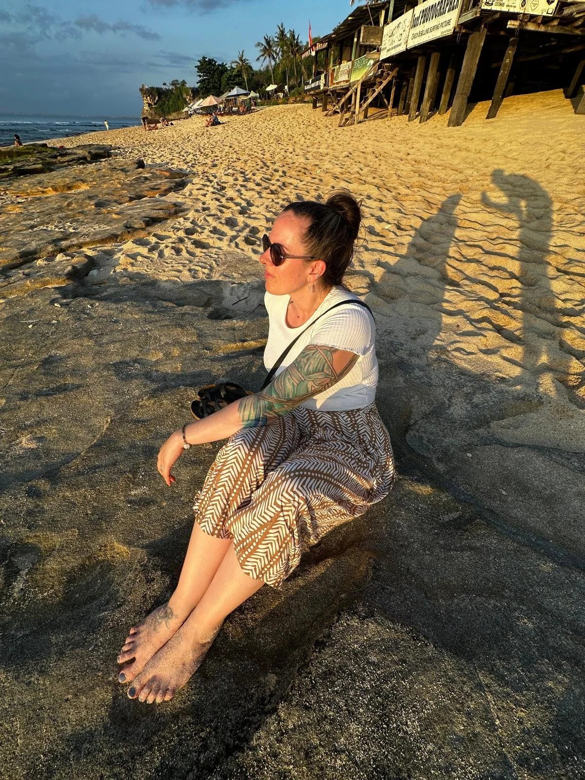 Eine Frau sitzt entspannt am Strand, ihre Füße im Sand, mit Blick auf das Meer, es ist Sonnenuntergang, Schatten sind sichtbar, im Hintergrund Zelte und Gebäude.