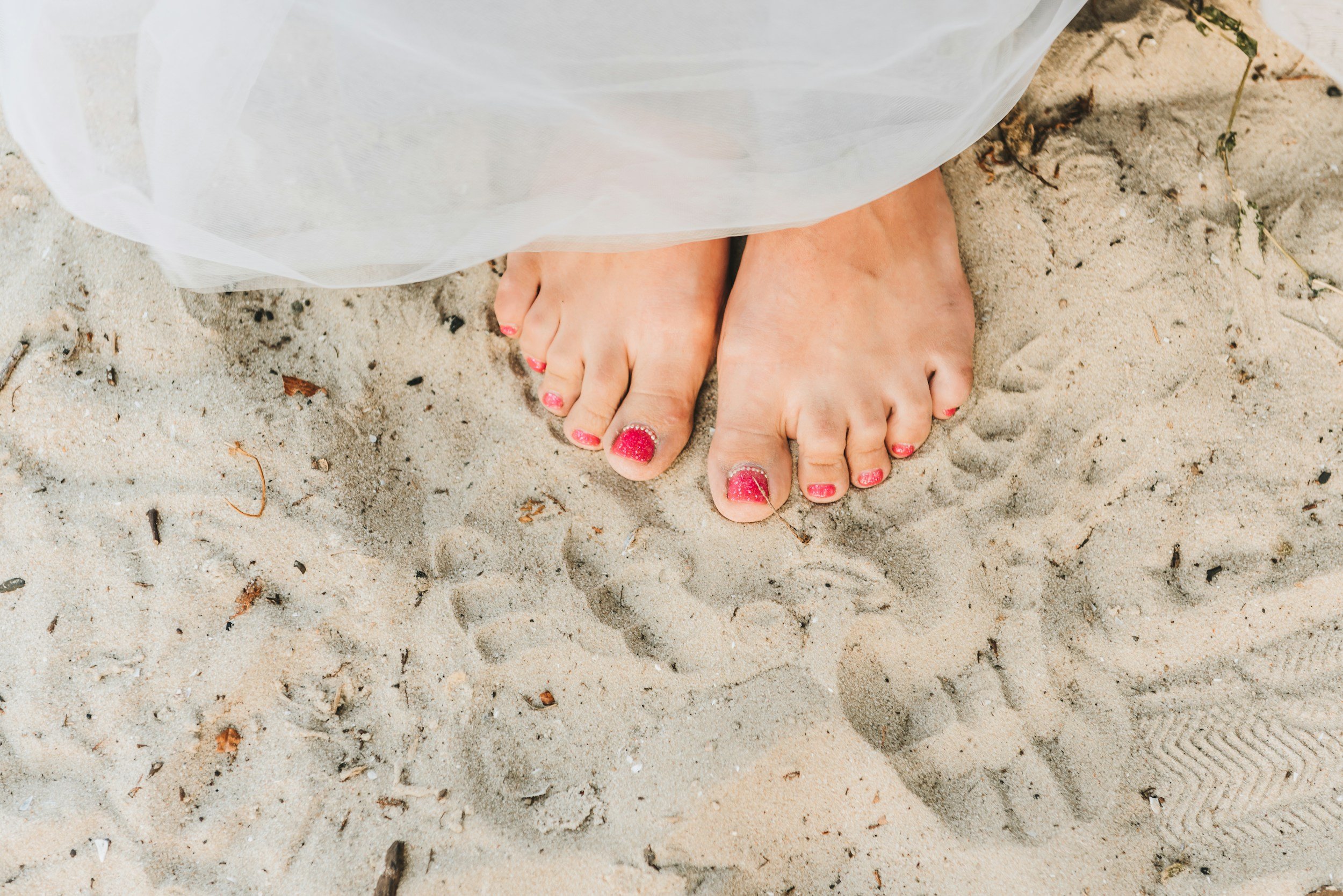 Nacktfüße mit roten, glitzernden Fußnägeln im Sand, teilweise von einem weißen Kleid bedeckt.