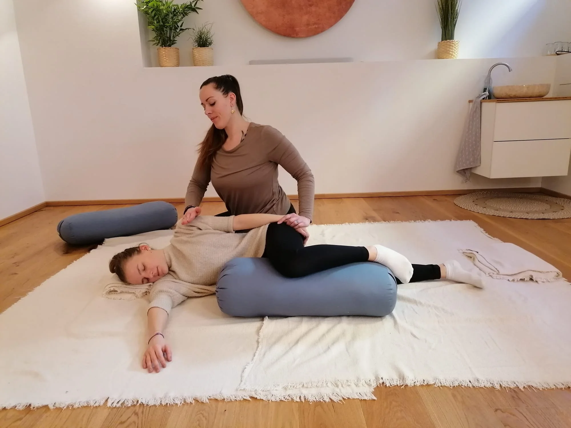 Eine Frau führt eine Yoga-Übung mit einem Kind aus, das auf einem weichen Teppich liegt, während sie in einer ruhigen Umgebung sitzt.