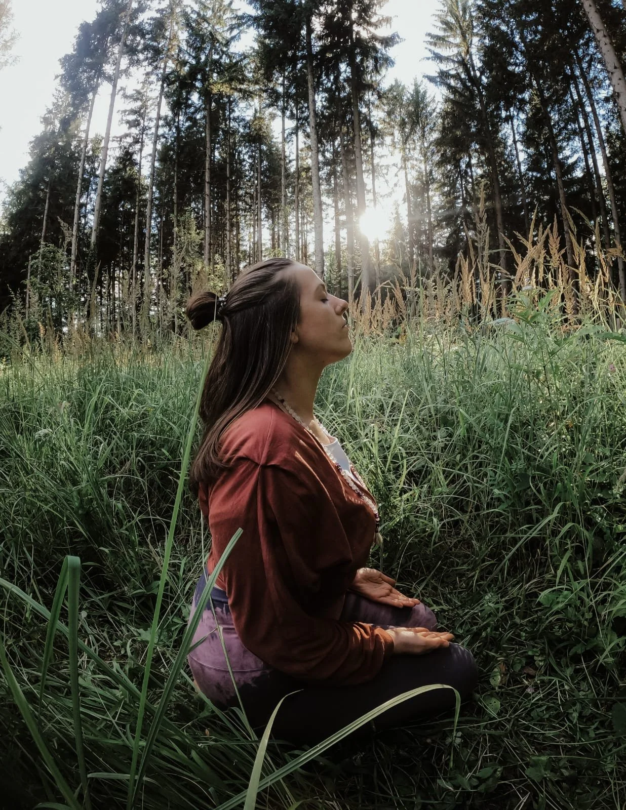 Eine Frau sitzt im Sitzen in einem grünen Wiesenfeld, umgeben von hohen Gräsern und hinter ihr ein Zaun. Die Sonne scheint durch die Bäume im Wald im Hintergrund, während sie in ruhiger Meditation mit geschlossenen Augen sitzt.