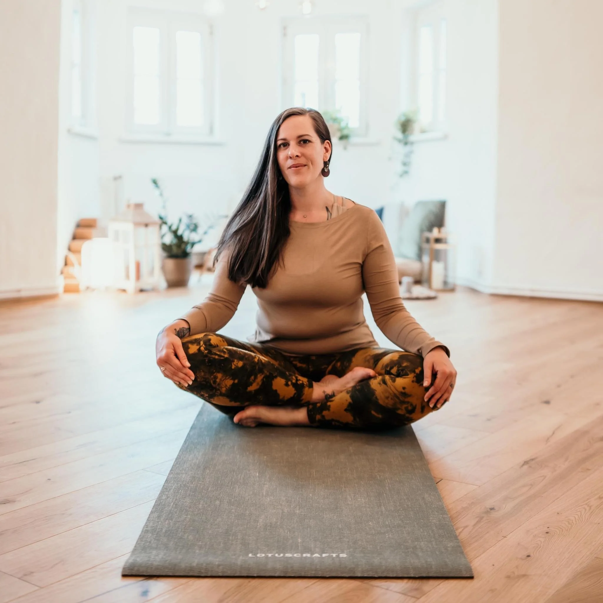 Eine Frau in bequemer Kleidung sitzt im Schneidersitz auf einem Yogamatte in einem hellen Raum mit großen Fenstern.