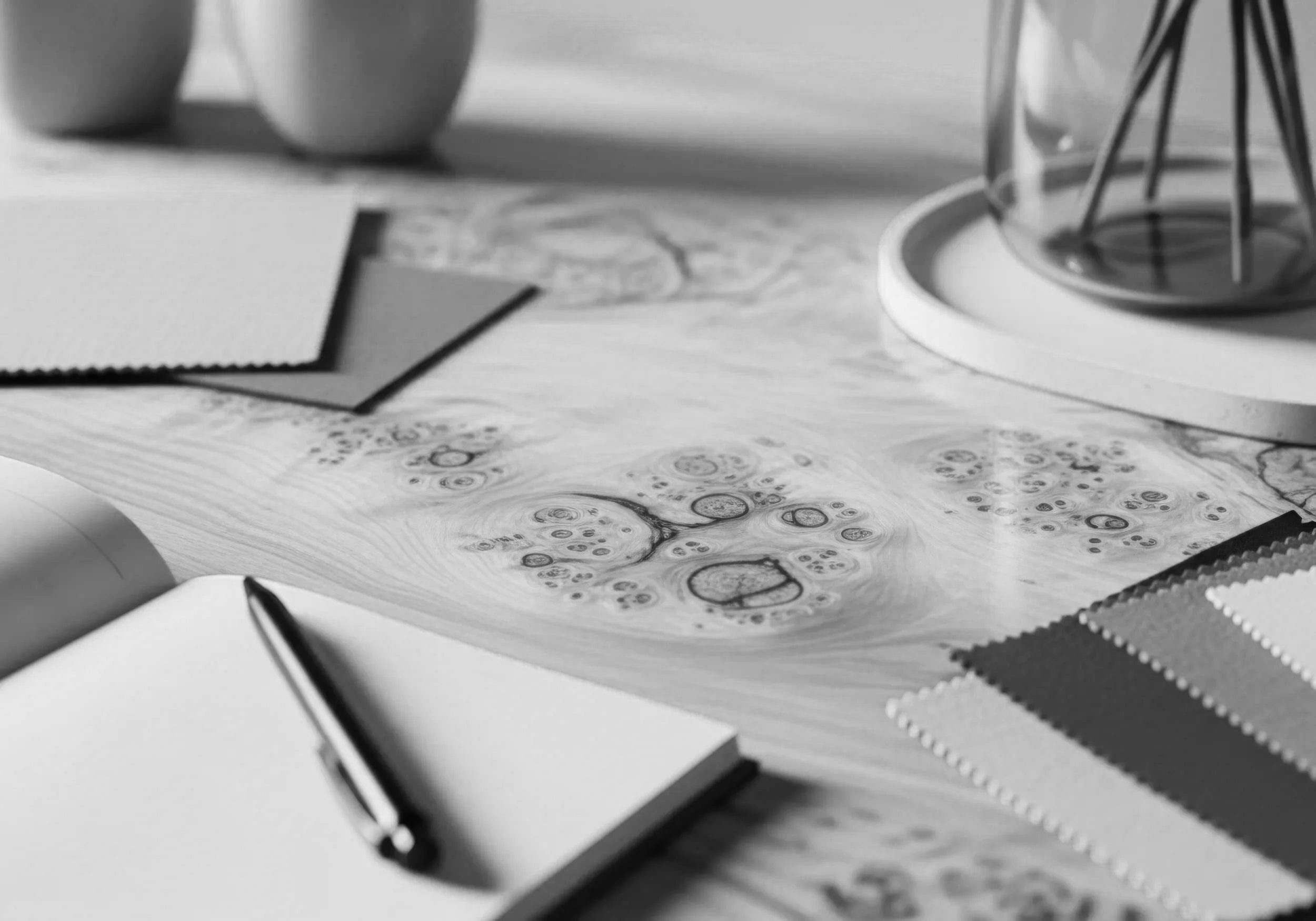 Black and white photo of notebooks, papers, and writing utensils on a desk with some objects in the background.