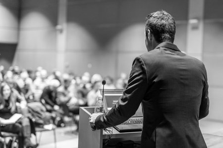 A man in a suit giving a presentation at a podium in front of an audience in a conference room.