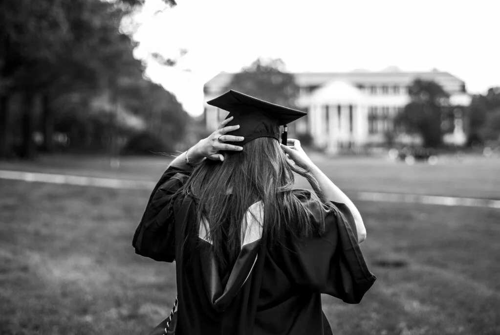 A woman in graduation gown and cap adjusting her cap on an outdoor campus.