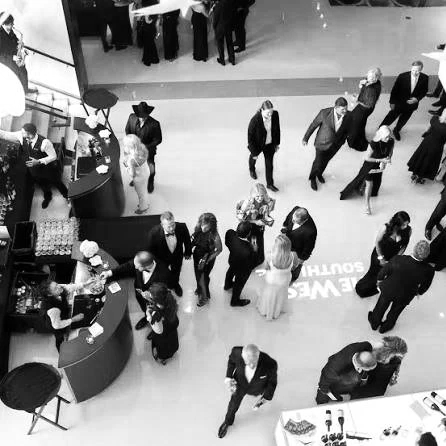 People gathered at an indoor event, mingling and talking around a bar and a table with drinks and food, in a large open space.