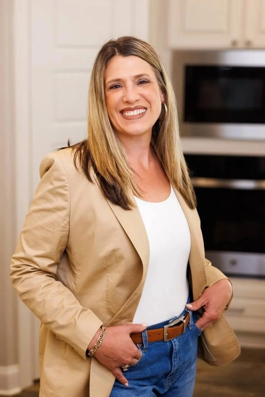 A woman with shoulder-length blond hair wearing a beige blazer, white top, and blue jeans, standing in a kitchen with a microwave in the background.