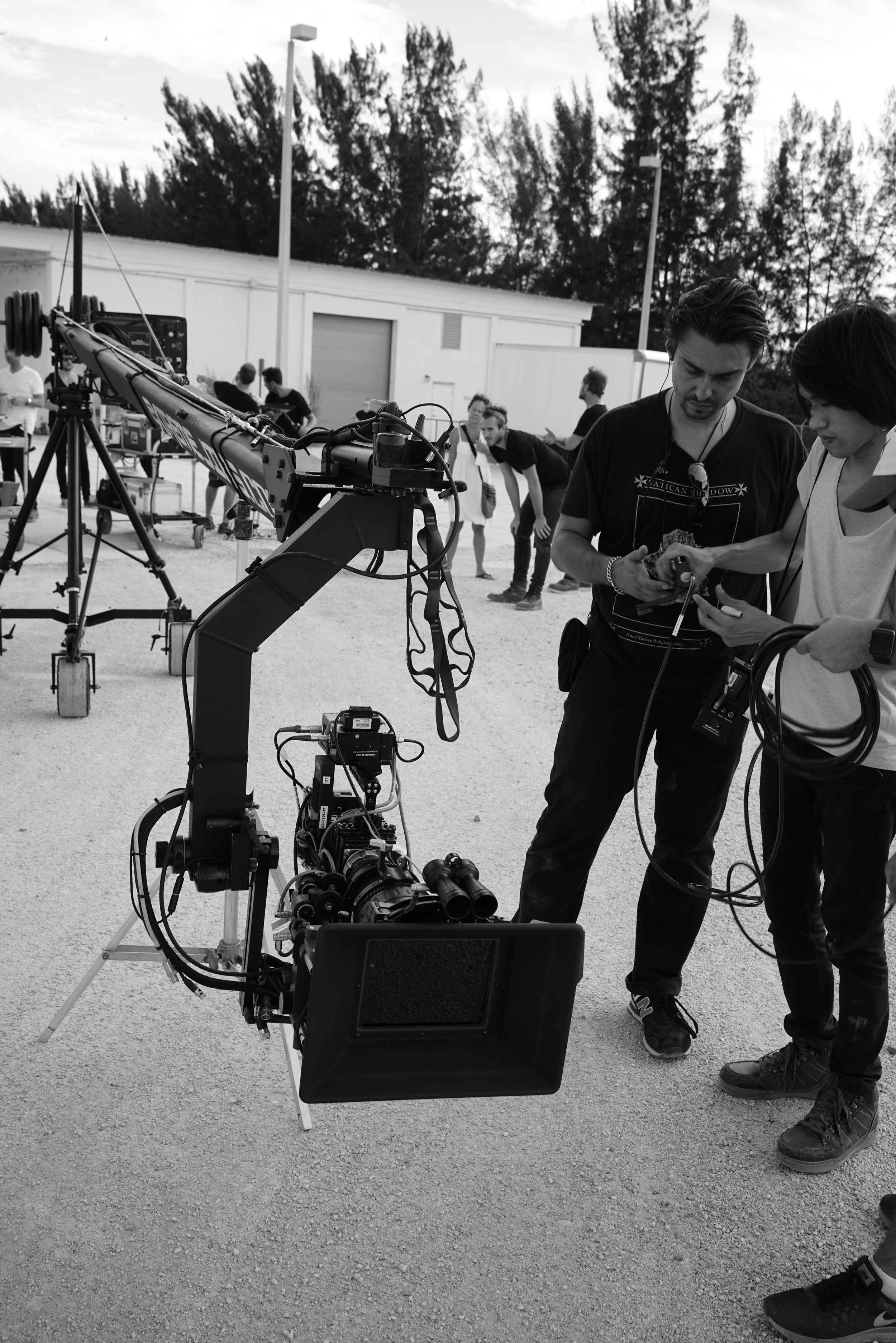 Behind the scenes of a film or video shoot outdoors. Crew members are working with camera and equipment setup, with a focus on two people examining a device or monitor. There are multiple crew members and equipment in the background, with trees and a building.